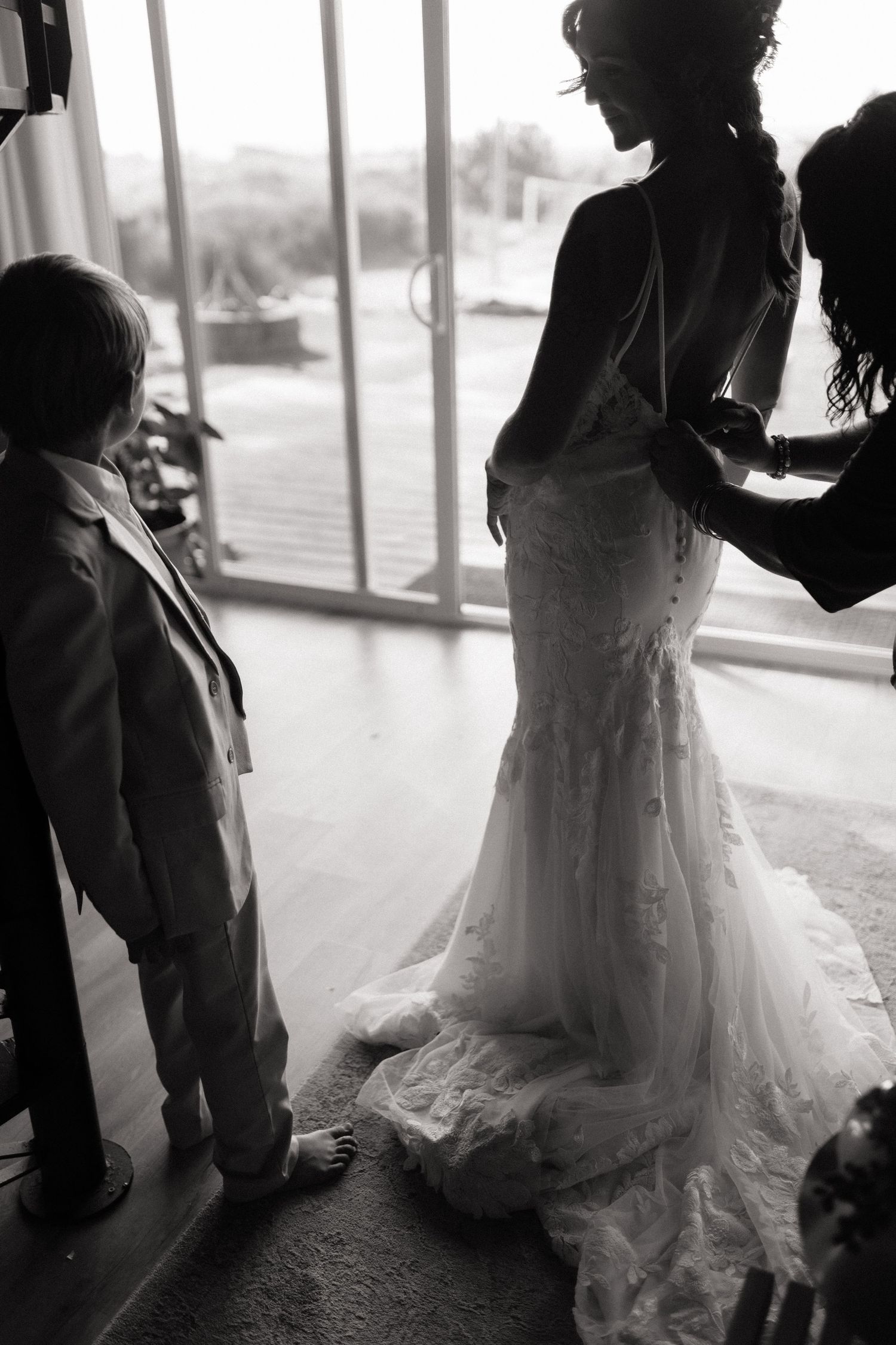 Black and white image of a young ring bearer watching a bride get her dress fastened.