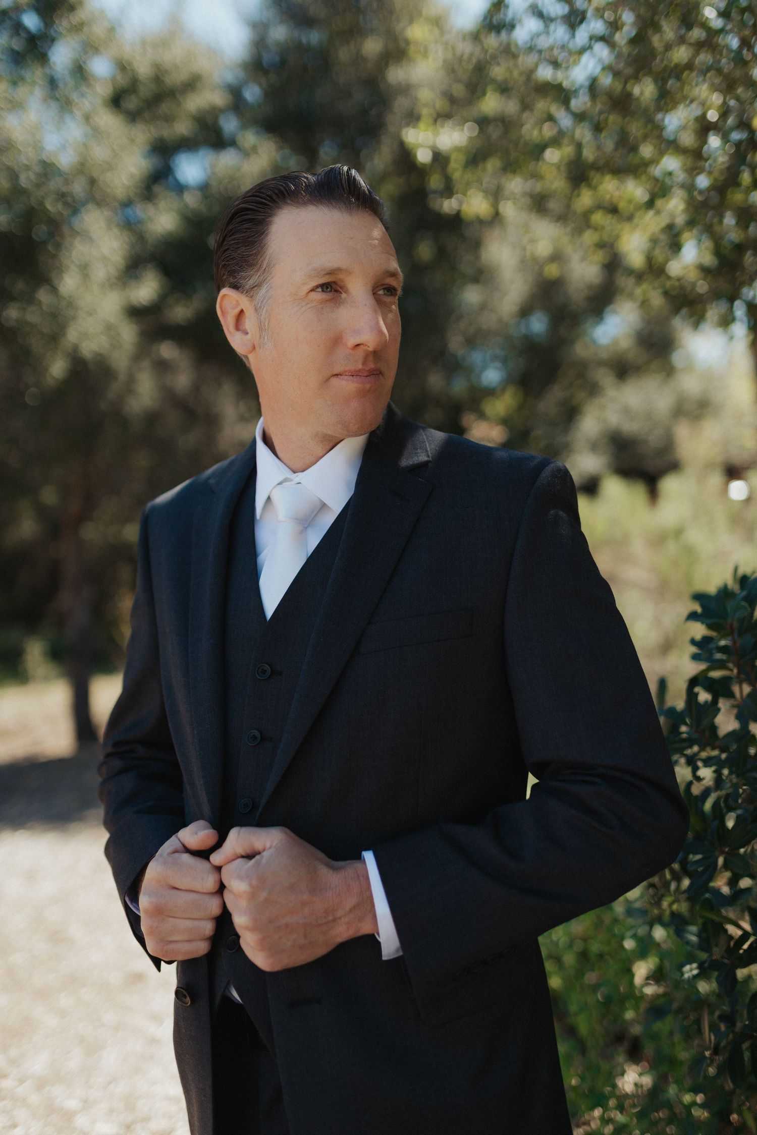A dapper groom in a black three-piece suit adjusts his jacket while standing outdoors among lush green trees.