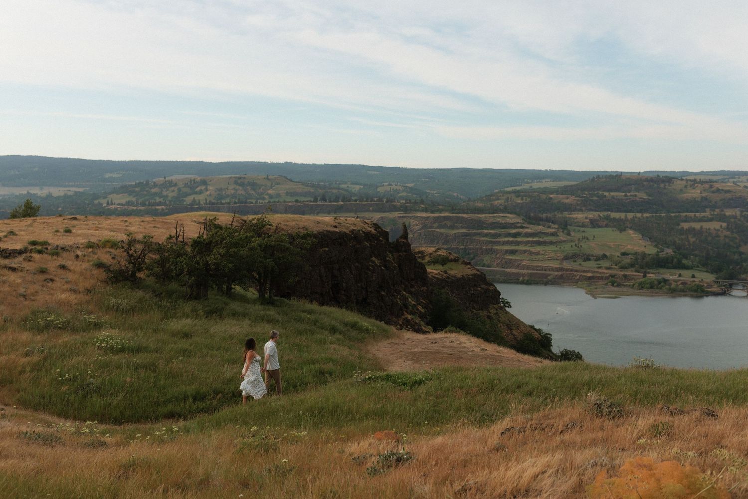 Couple strolling along a grassy cliff overlooking a scenic river valley with dramatic landscape views.