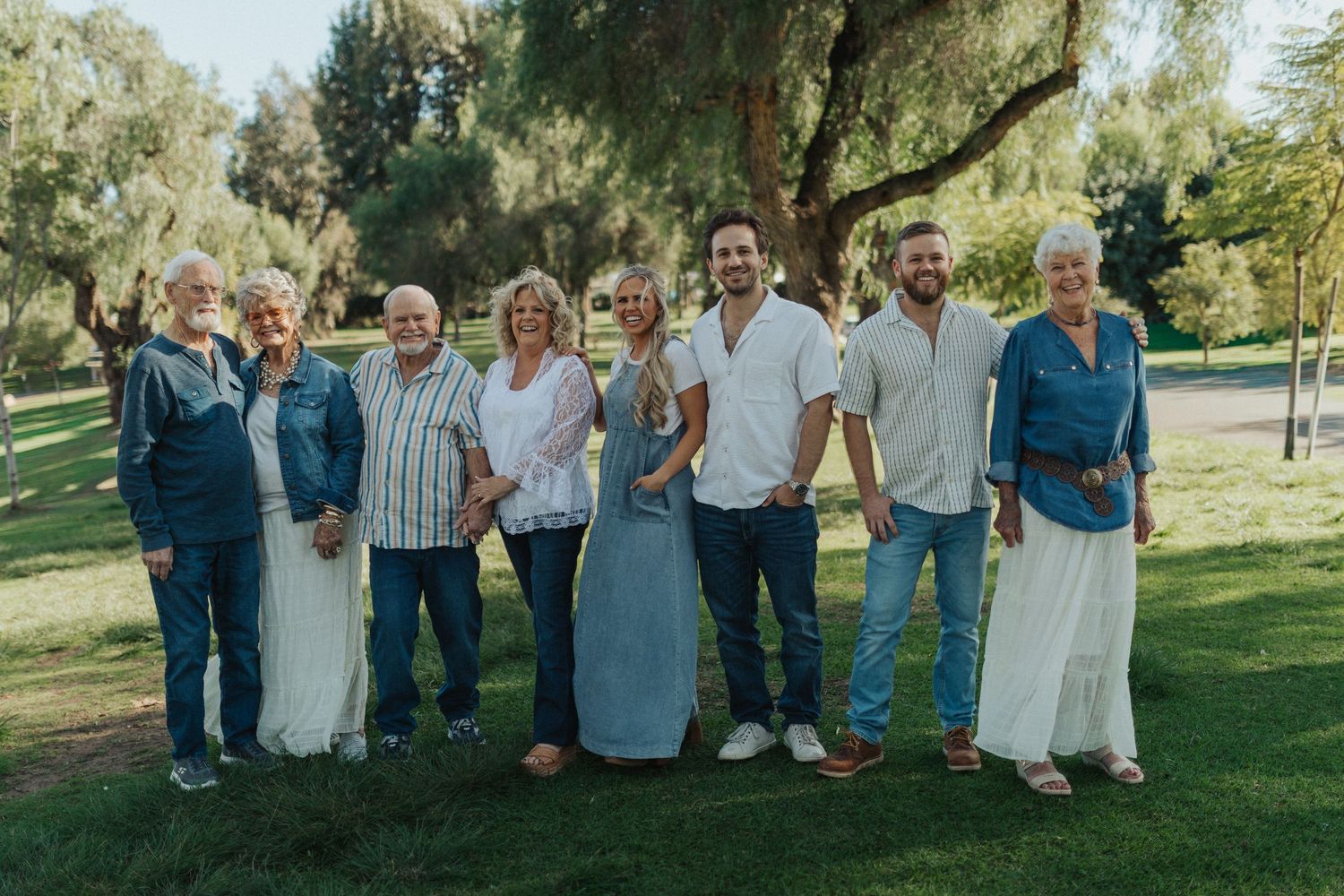 A multigenerational family of eight smiles together on green grass beneath shady trees in a park.