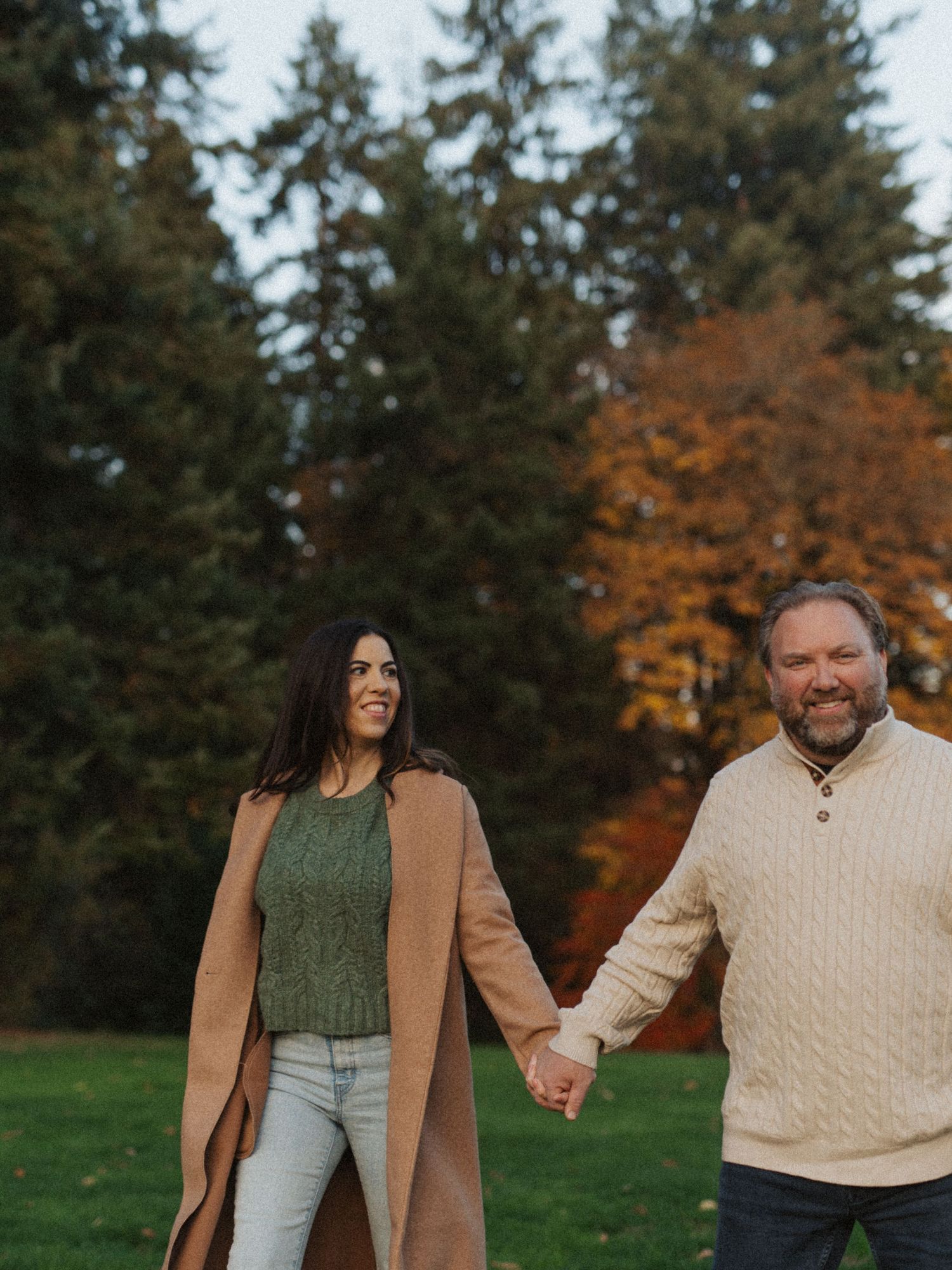 A smiling couple holds hands while walking through a park with vibrant fall foliage in the background.