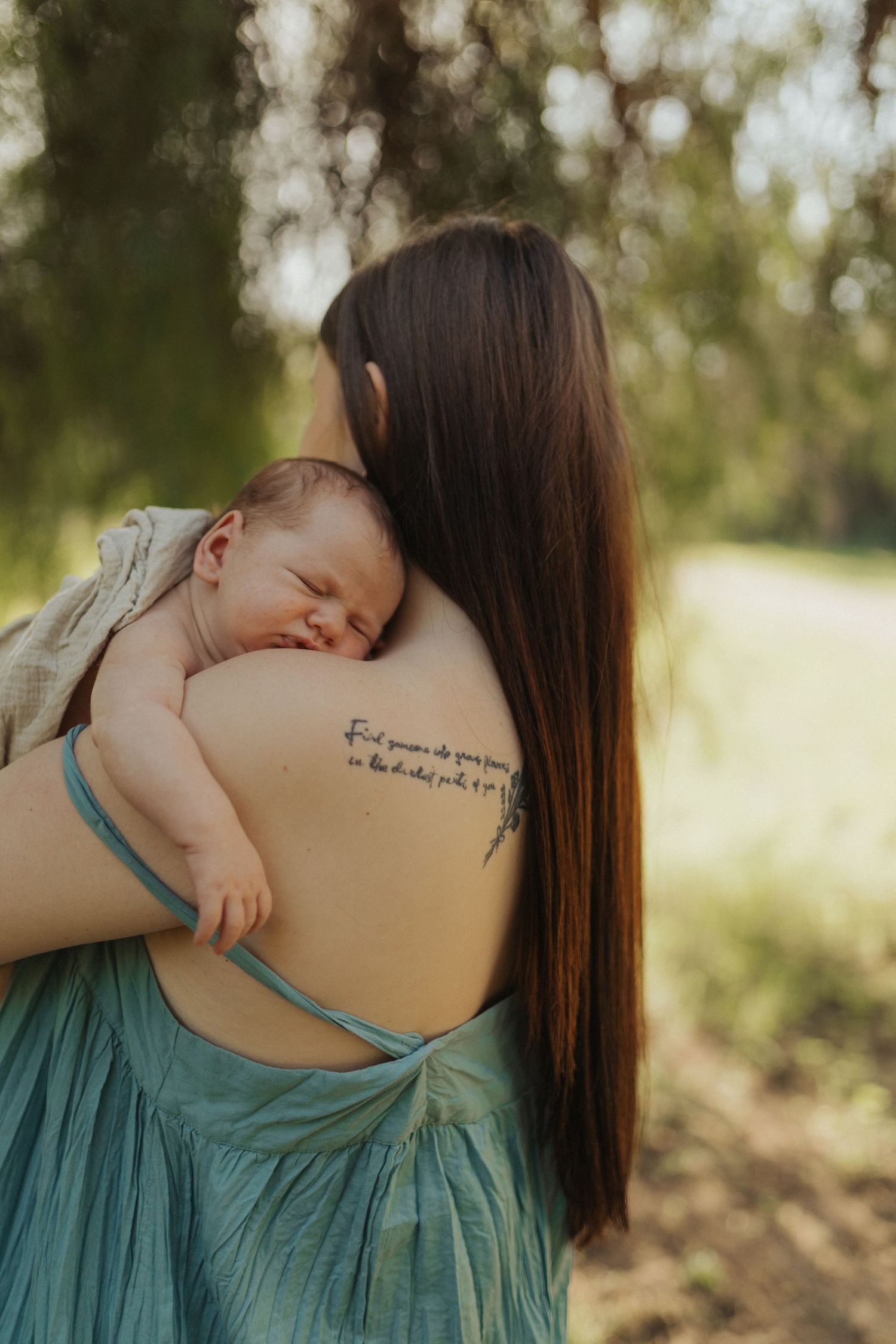 Mother with back tattoo holding sleeping newborn over shoulder in lush green outdoor setting.