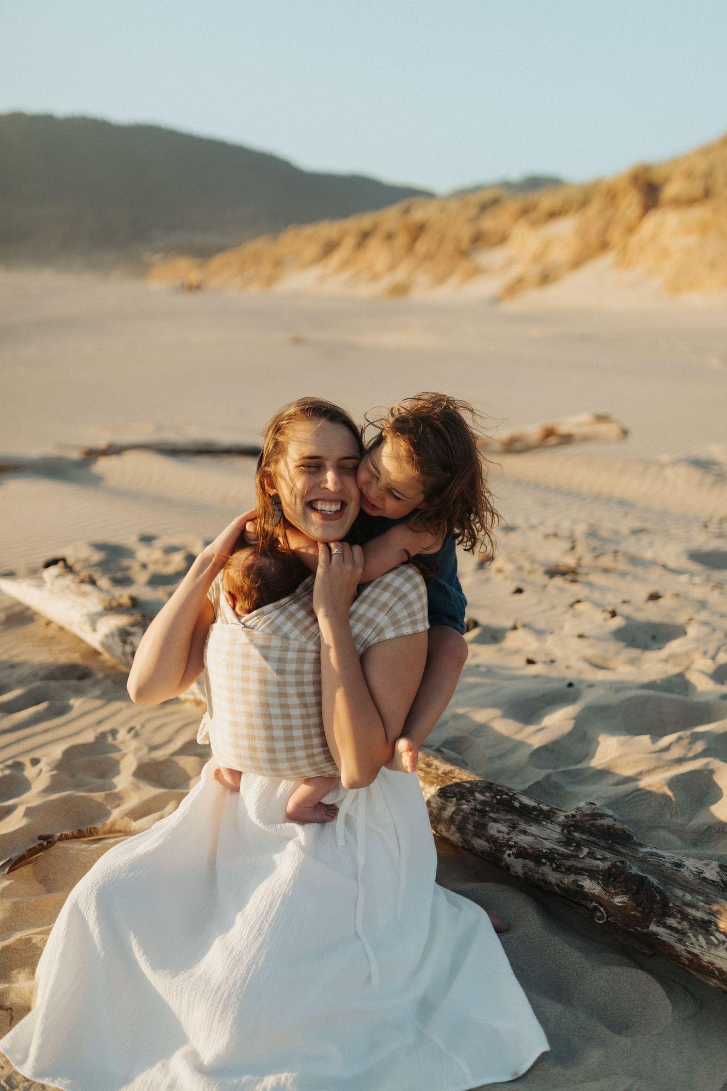 Mother laughing joyfully as young daughter hugs her from behind while sitting on sandy beach.