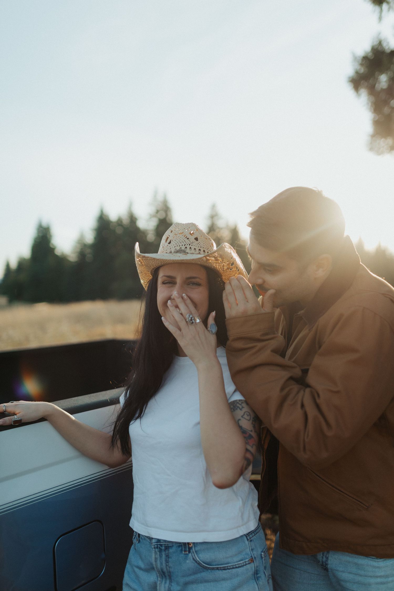 Laughing woman in cowboy hat and white tee with man in brown jacket beside a vintage pickup truck.