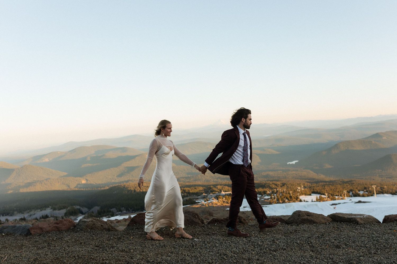 Bride and groom hold hands walking on mountain summit with scenic valley views at golden hour wedding. Mt. Hood Silcox Hut Wedding.