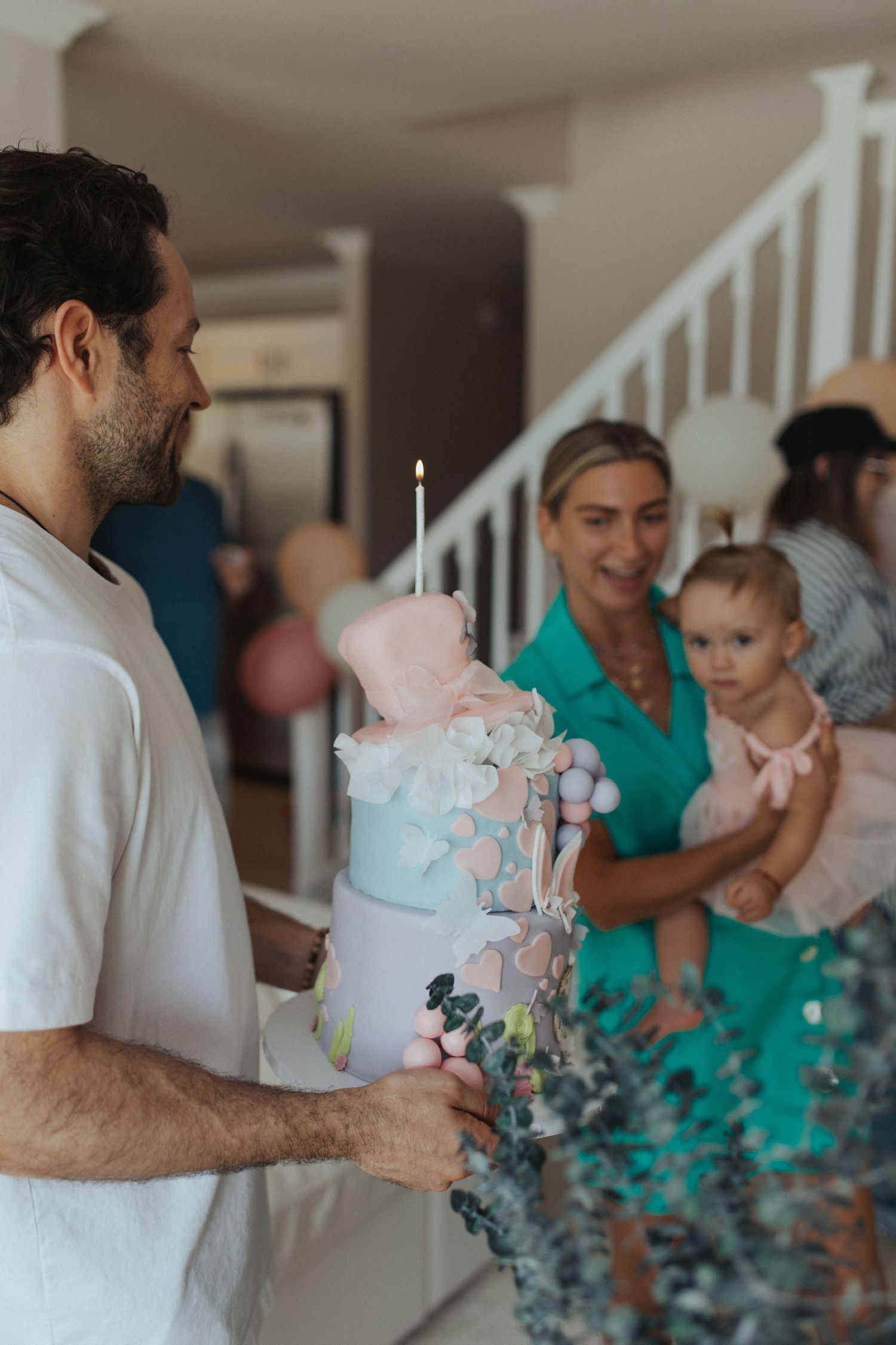 Man in white shirt holds lit candle birthday cake while woman holds baby at celebration.