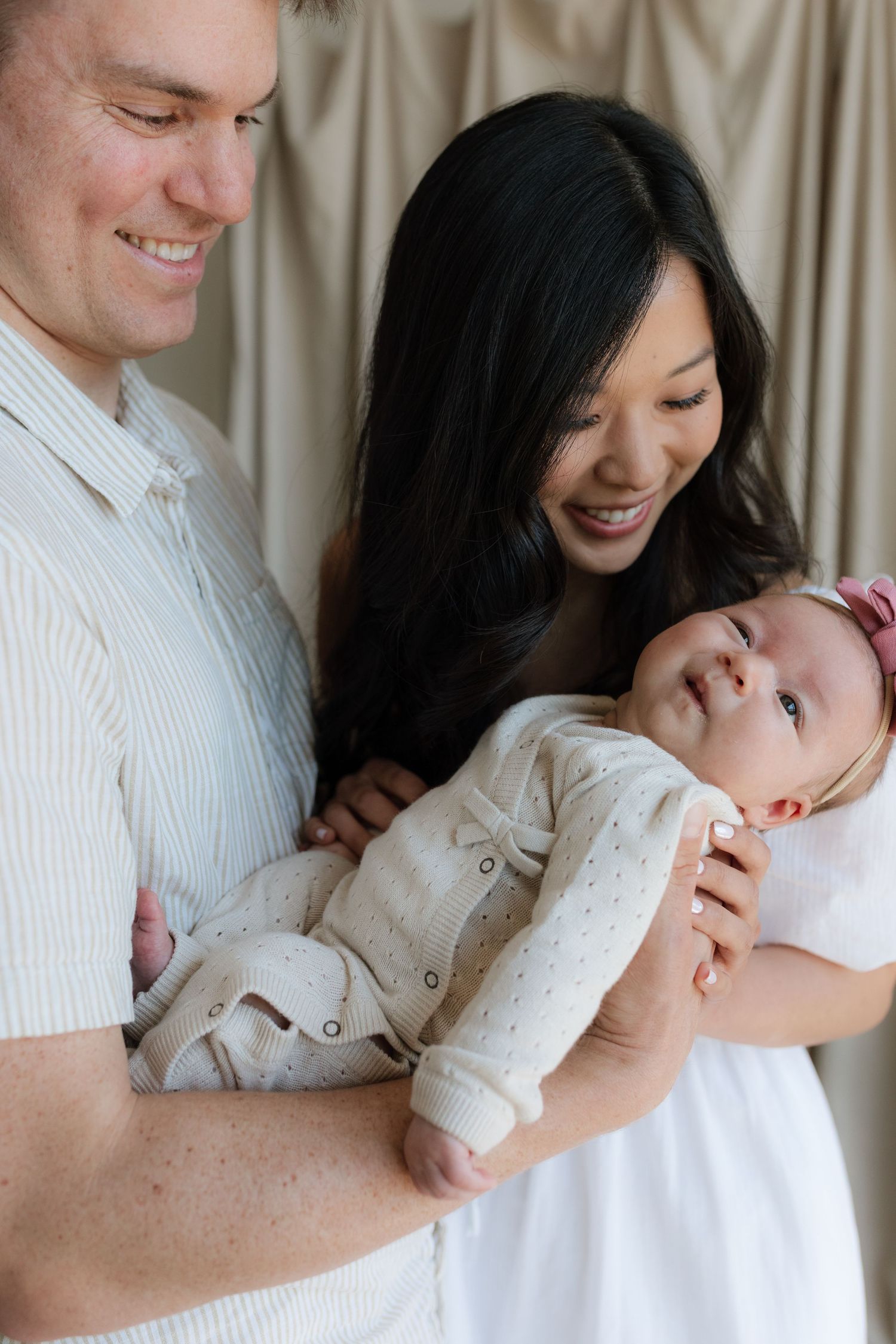 Smiling parents hold their wide-eyed newborn baby dressed in a cream knit outfit.