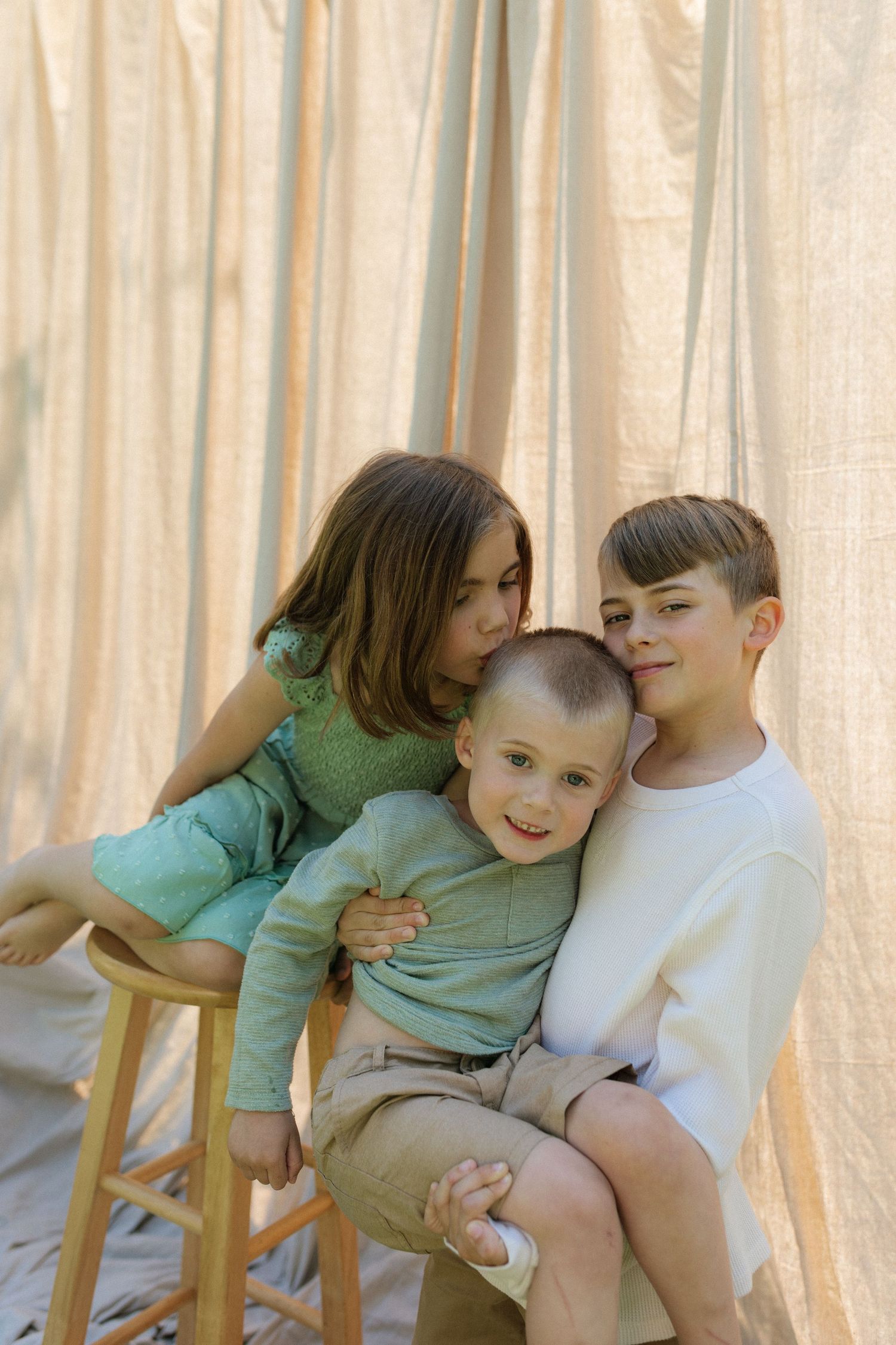 Three siblings share a warm embrace on a stool against a beige linen curtain backdrop outdoors.