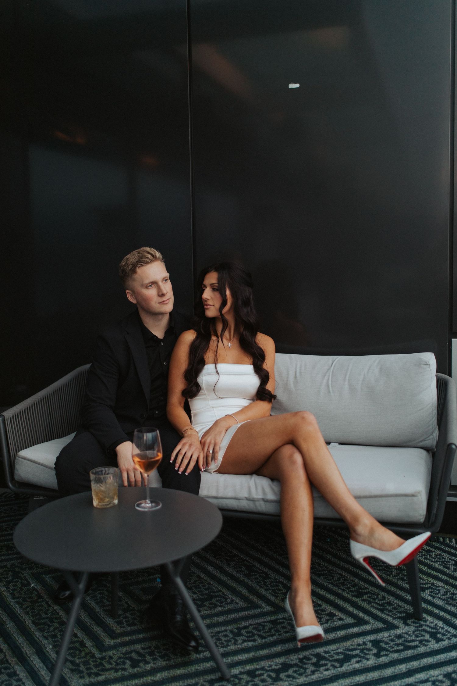 Elegant couple relaxing on outdoor lounge sofa with drinks, woman in white mini dress wearing red sole heels.