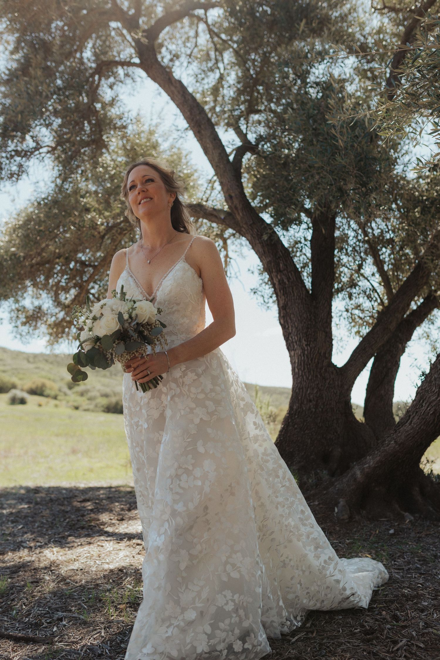 Bride in floral lace gown holds white bouquet while gazing upward beneath a rustic olive tree.