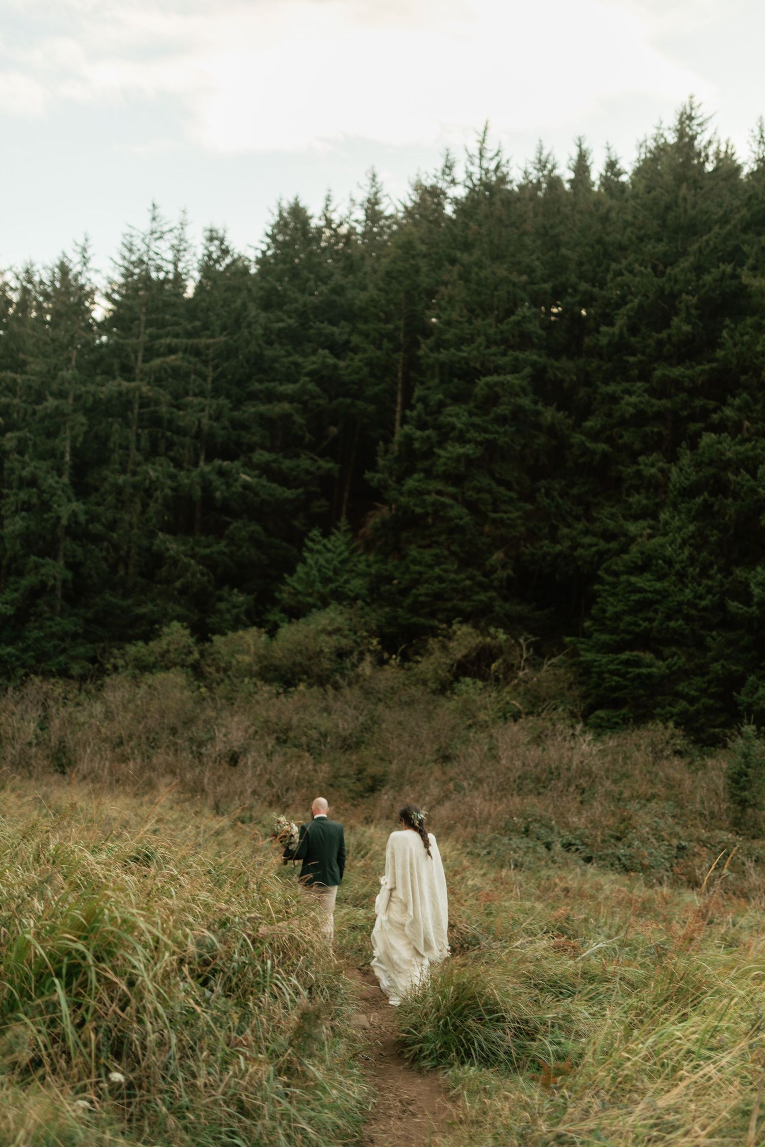 Bride and groom walking hand in hand through tall grass toward a pine forest.