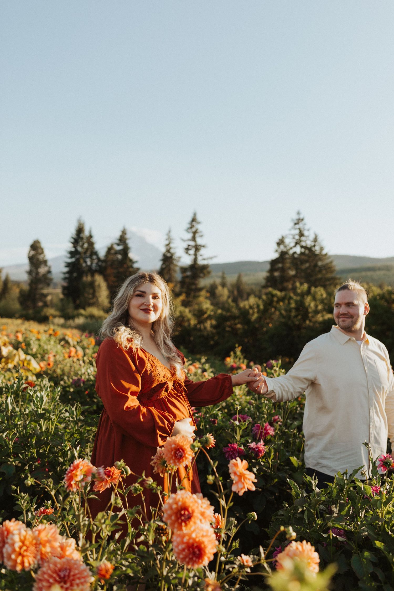 Pregnant woman in red dress holds partners hand in colorful dahlia field with mountain view.