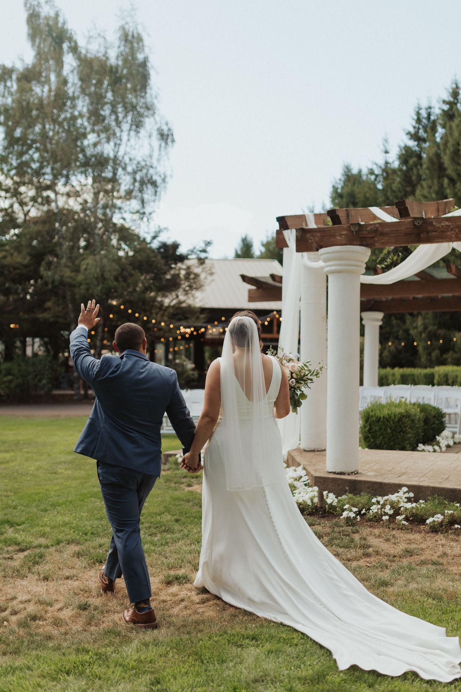 Bride and groom walk hand-in-hand toward a pergola adorned with string lights at an outdoor wedding venue.