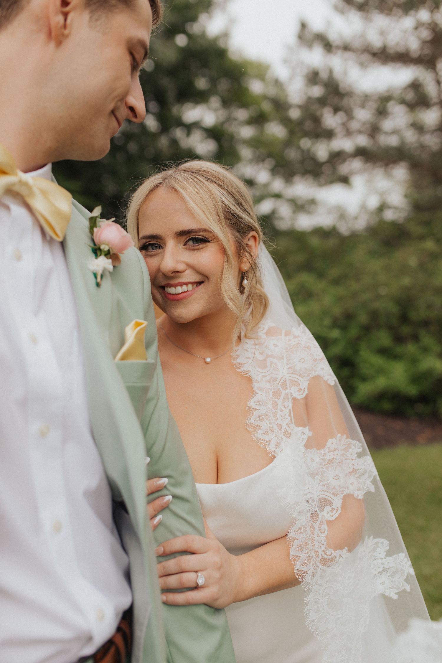 Smiling bride in lace veil holds grooms mint suit jacket arm during outdoor wedding portraits.