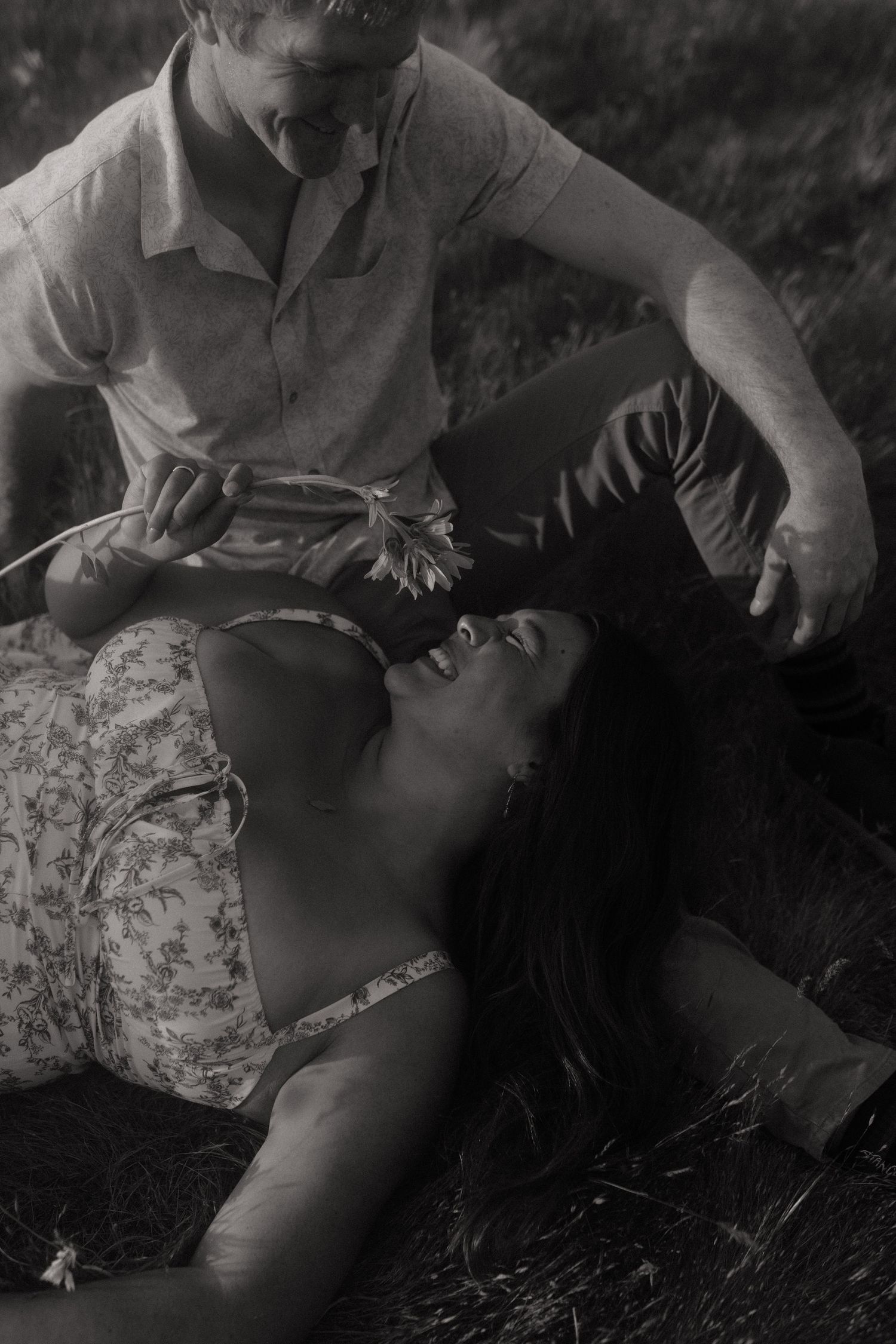 Black and white photo of a couple laughing together as the woman lies in the grass.