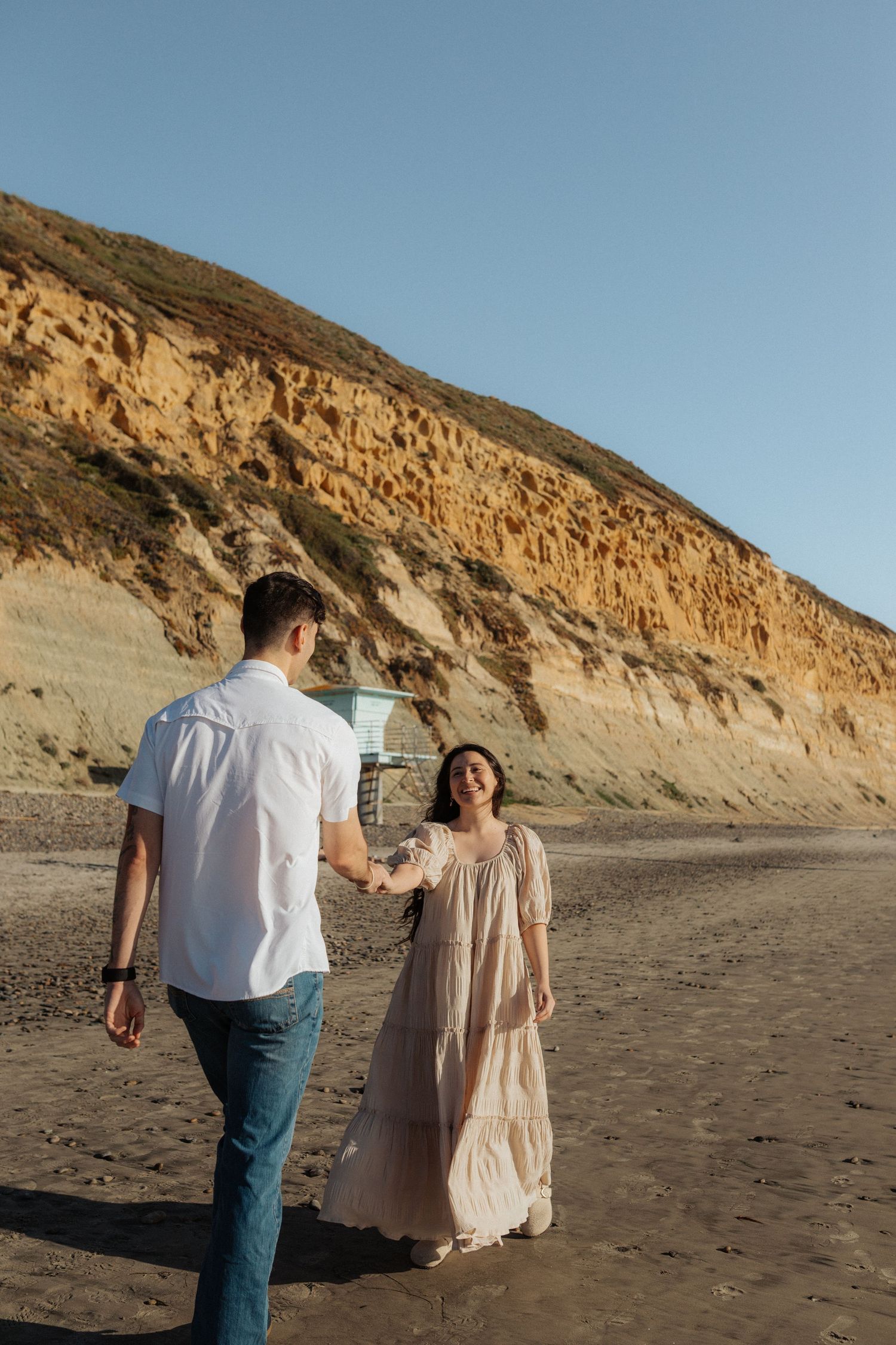 Couple holding hands on a sandy beach with dramatic golden cliffs and a lifeguard tower in the background.