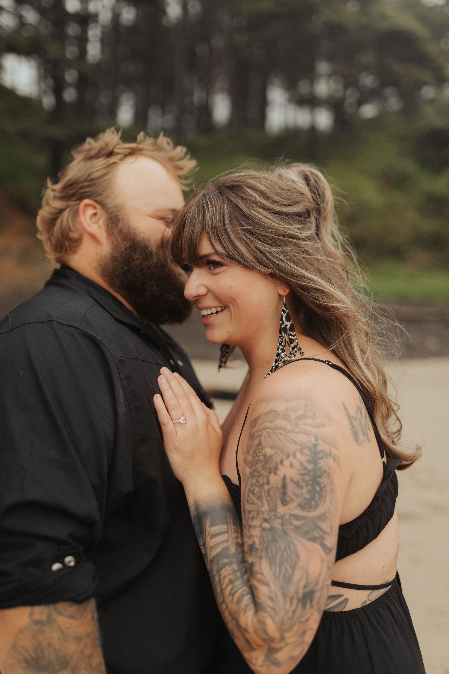 Smiling tattooed couple in black outfits sharing an intimate moment on a beach.