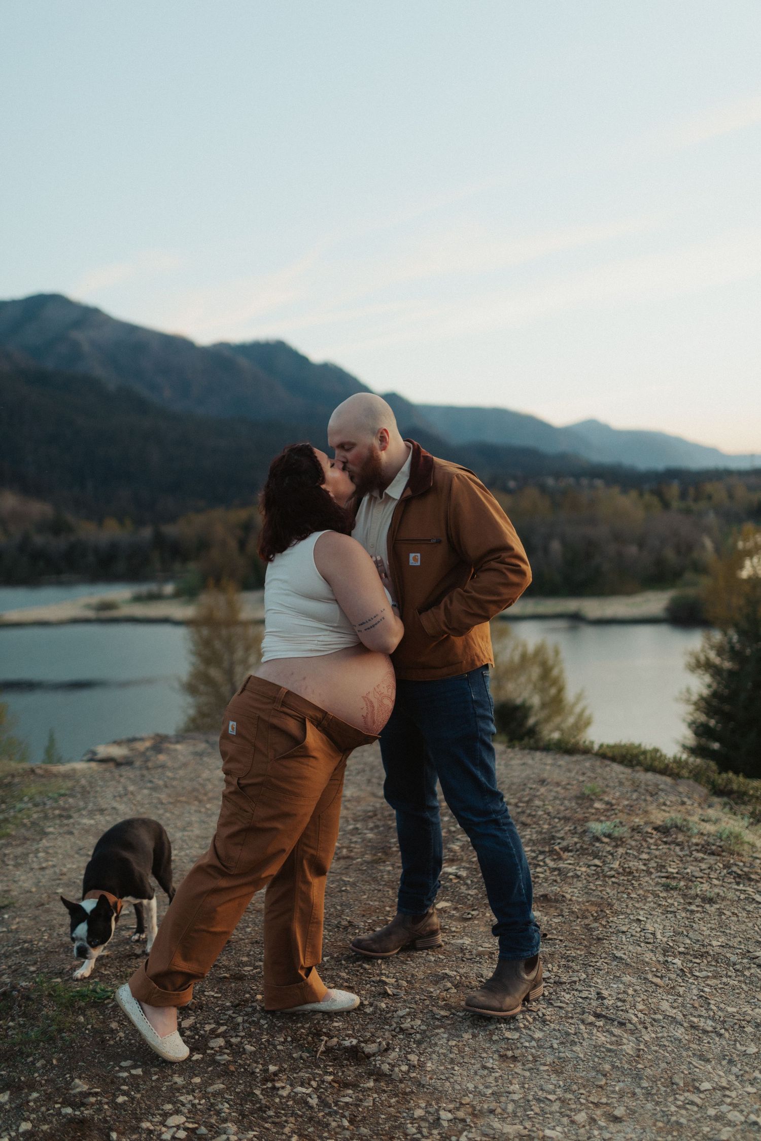 Pregnant couple kissing on a rocky overlook with a Boston Terrier and mountain lake views behind them.