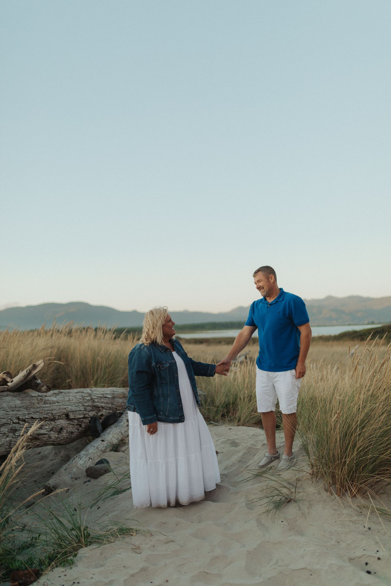 Couple holding hands on sandy beach dunes with tall grass and mountains in background at sunset.