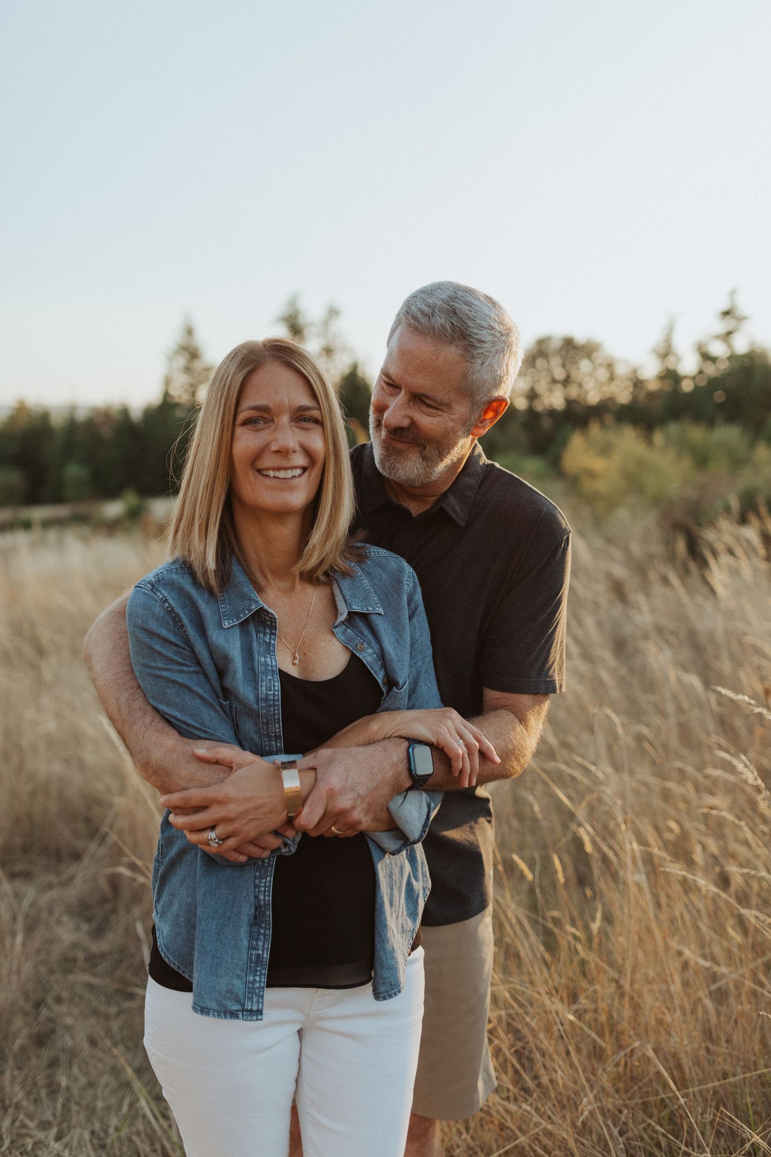 Man hugging woman from behind in a golden grass field at sunset.