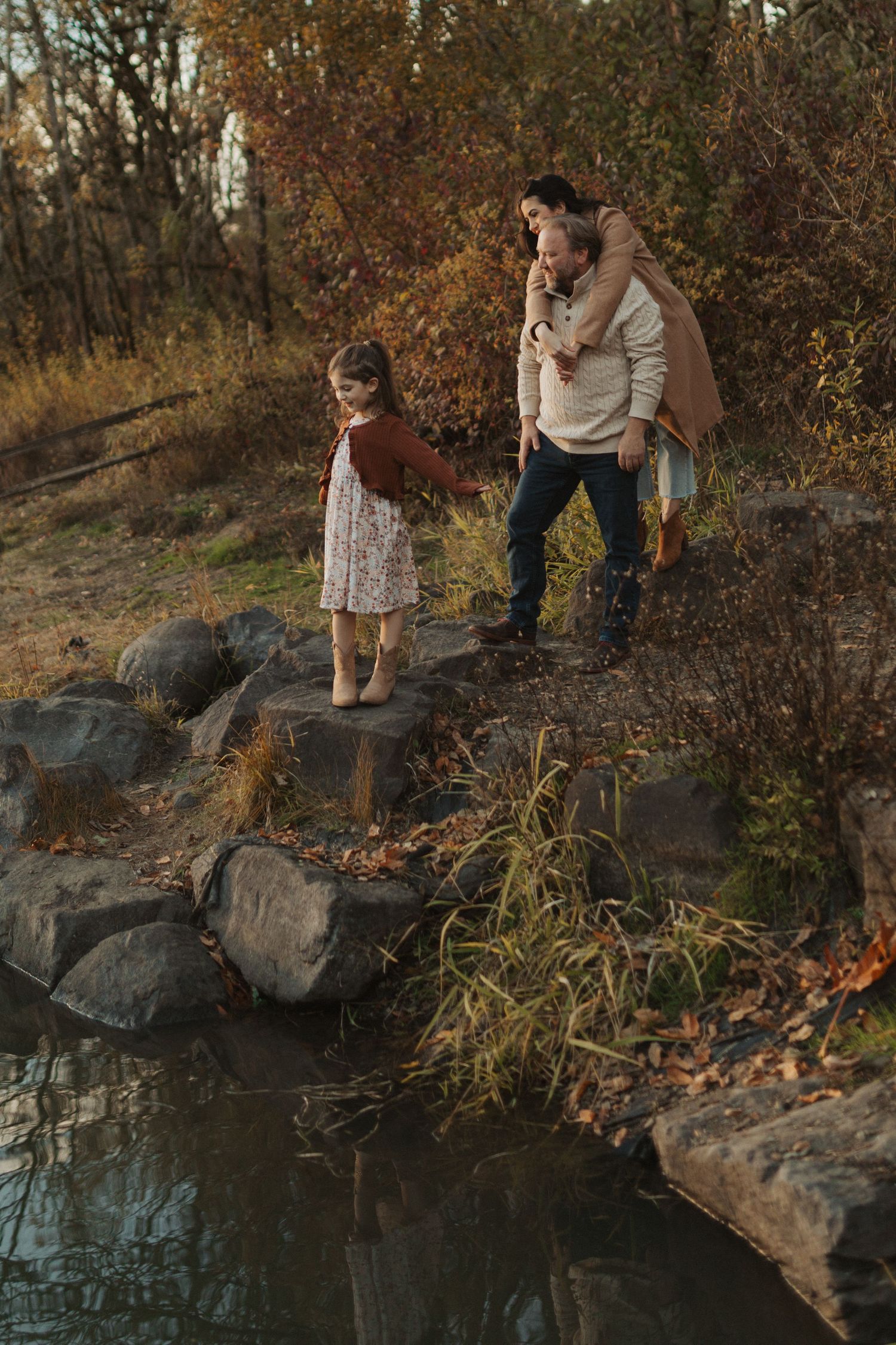 A family of three explores rocky terrain beside a pond surrounded by colorful autumn foliage.