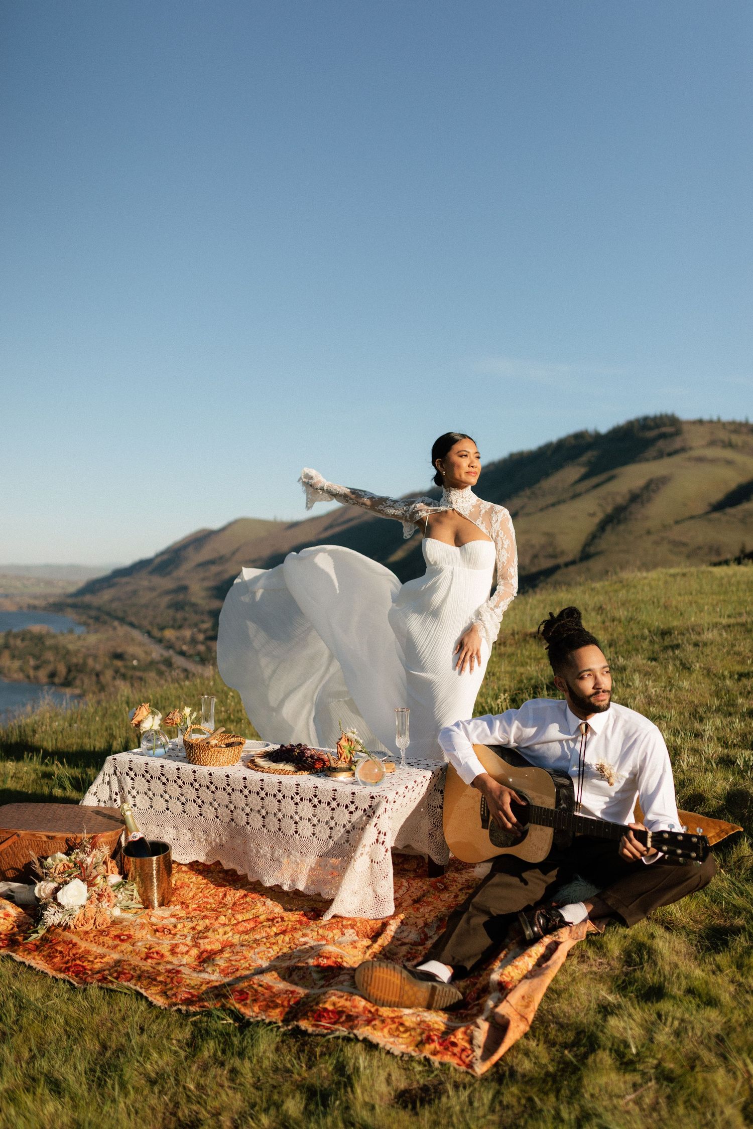 Bride in flowing white lace gown stands as groom plays guitar during a boho outdoor picnic with mountain views.