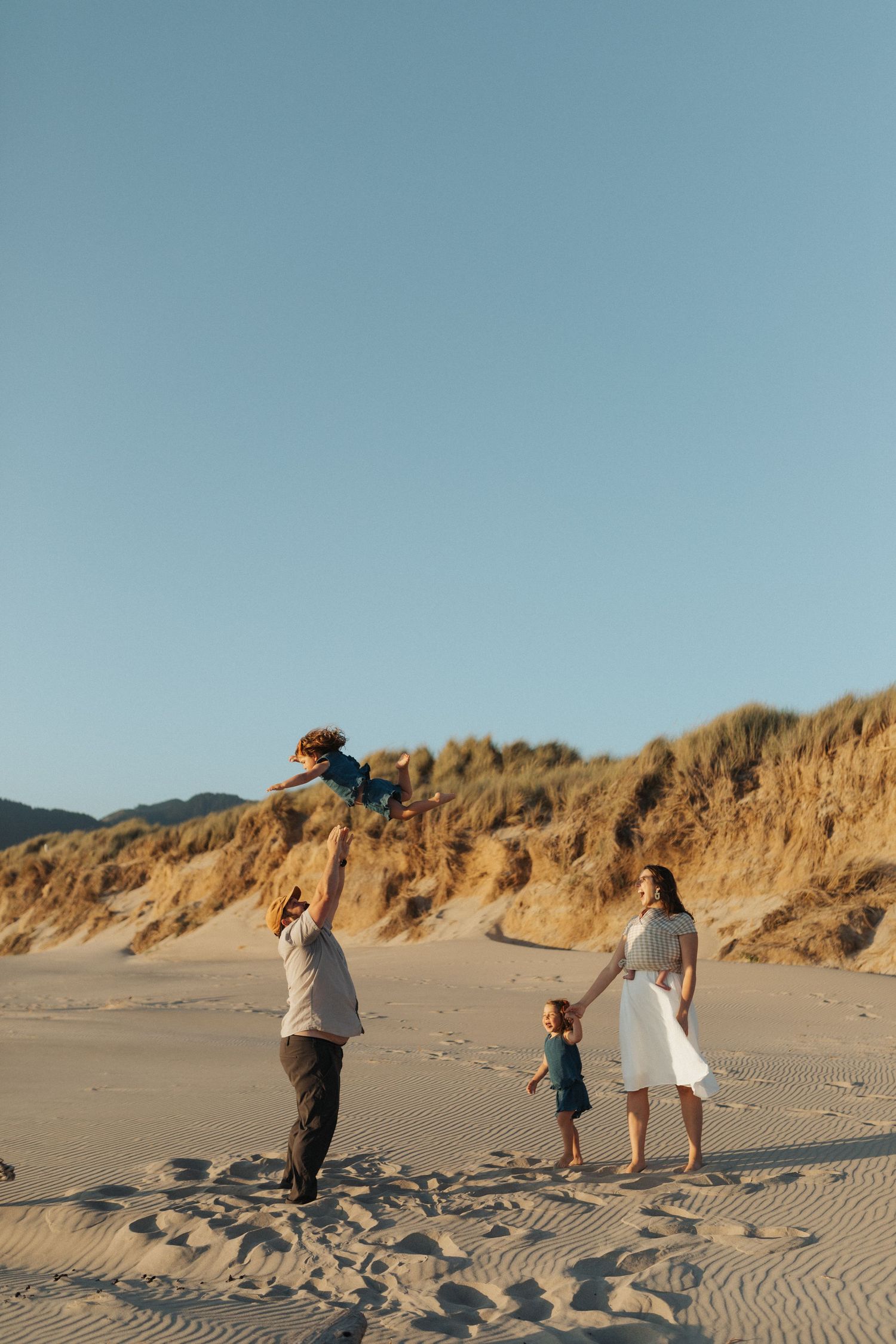 Family of four playing on sandy beach dunes, father tossing child in the air at sunset.