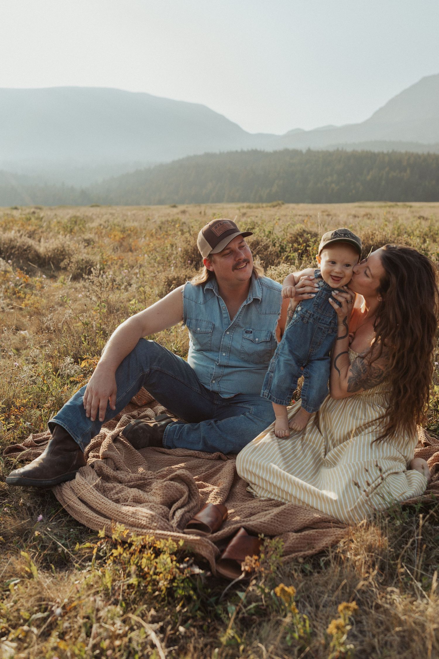 Happy family with baby sitting on blanket in scenic mountain meadow during golden hour outdoor photo session.