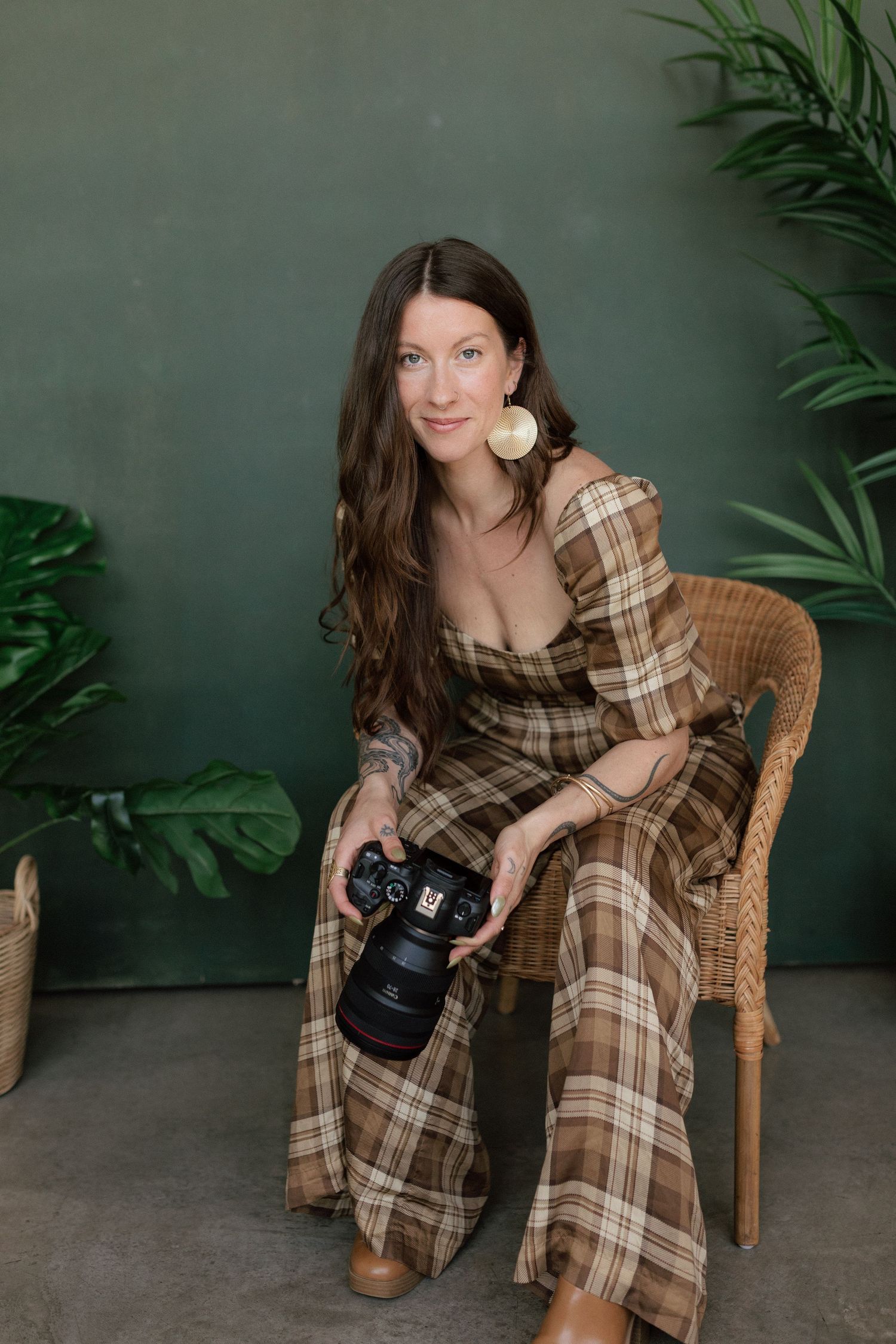 Female wedding photographer in plaid jumpsuit sitting in wicker chair holding Canon camera indoors.