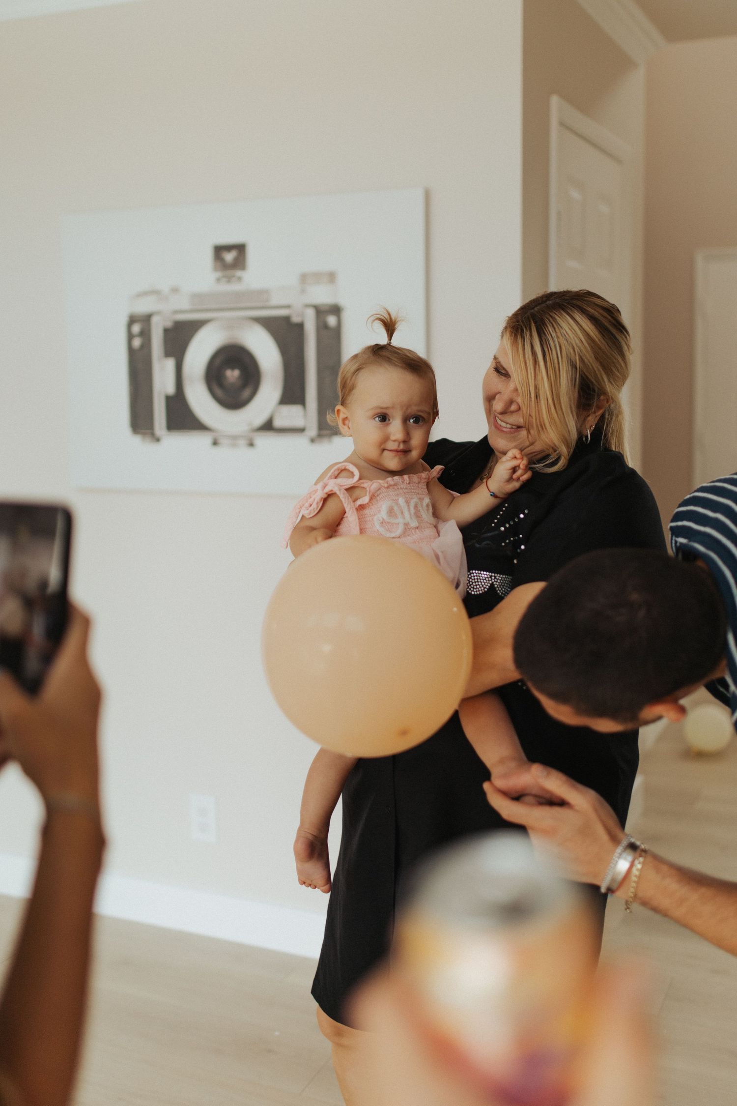 Woman holds baby girl in pink dress with balloon indoors.