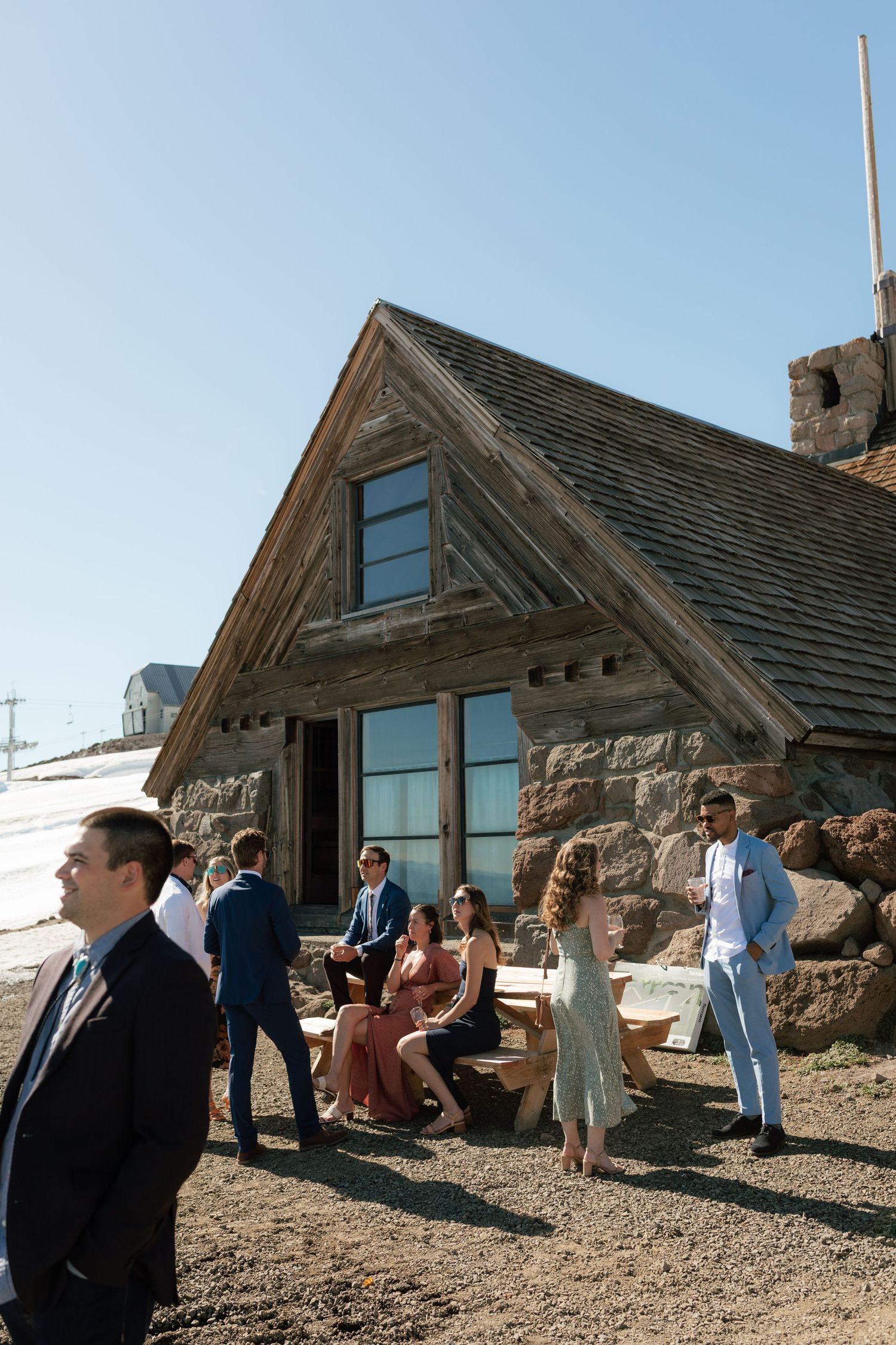 Wedding guests mingle outside rustic stone mountain lodge on sunny clear day.