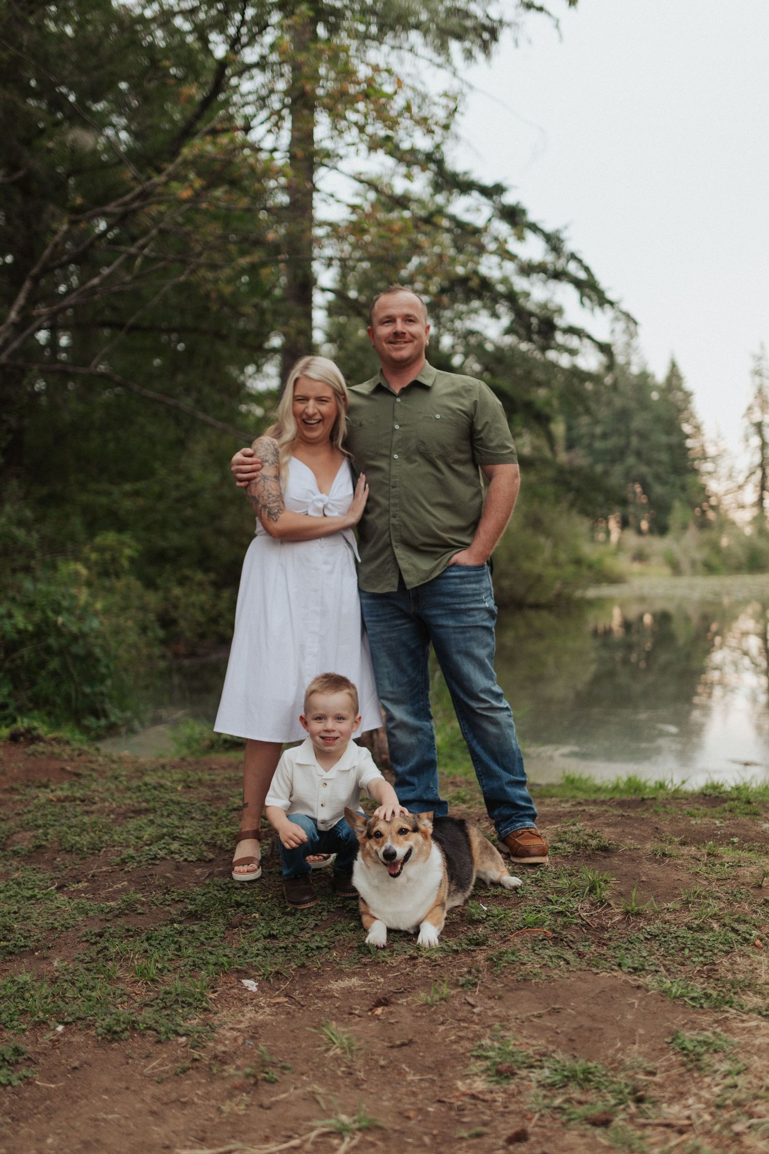 Smiling family portrait with young boy and corgi dog beside a calm woodland river.
