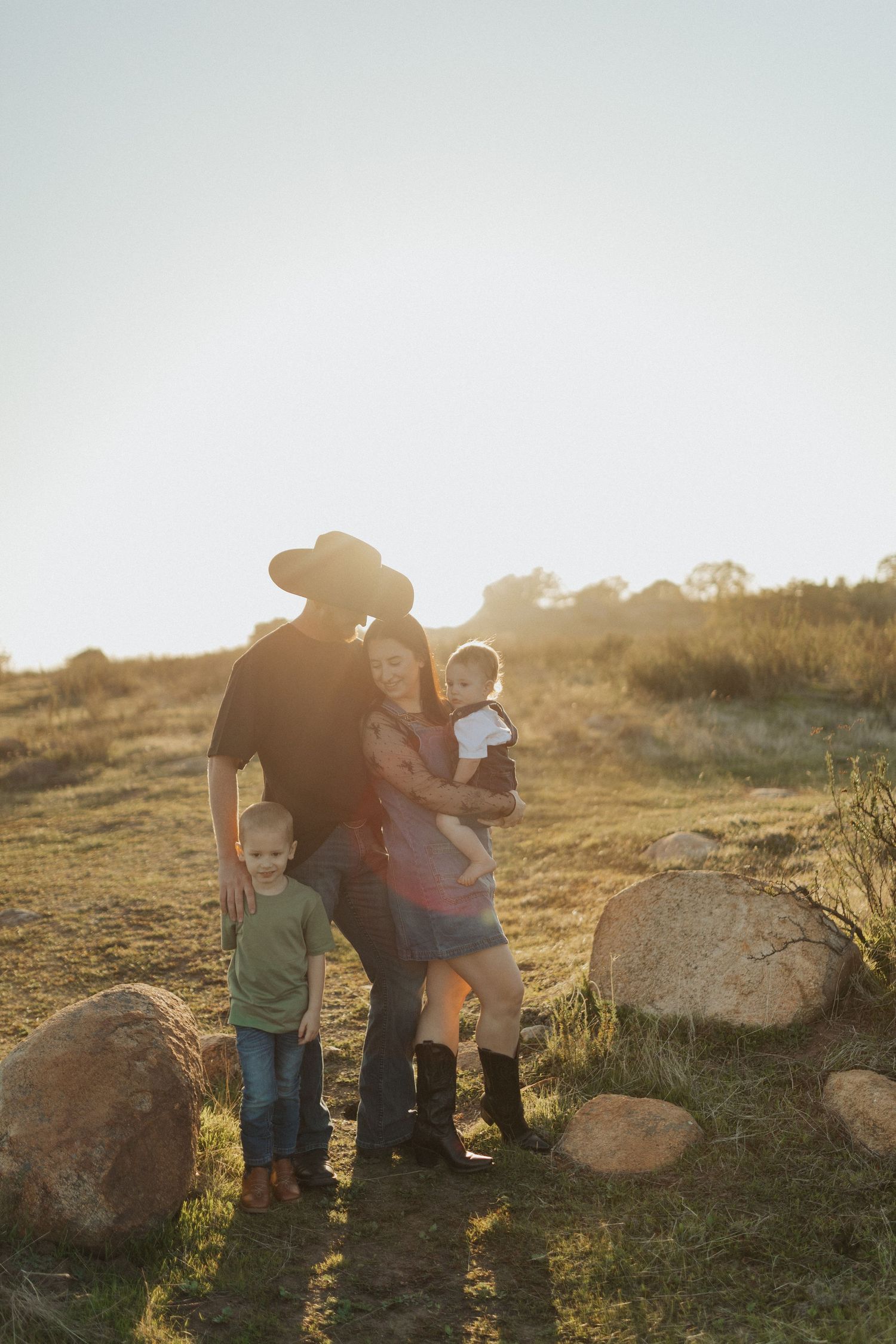 Family of four embracing in golden sunlit field with large rocks, father wearing black cowboy hat.