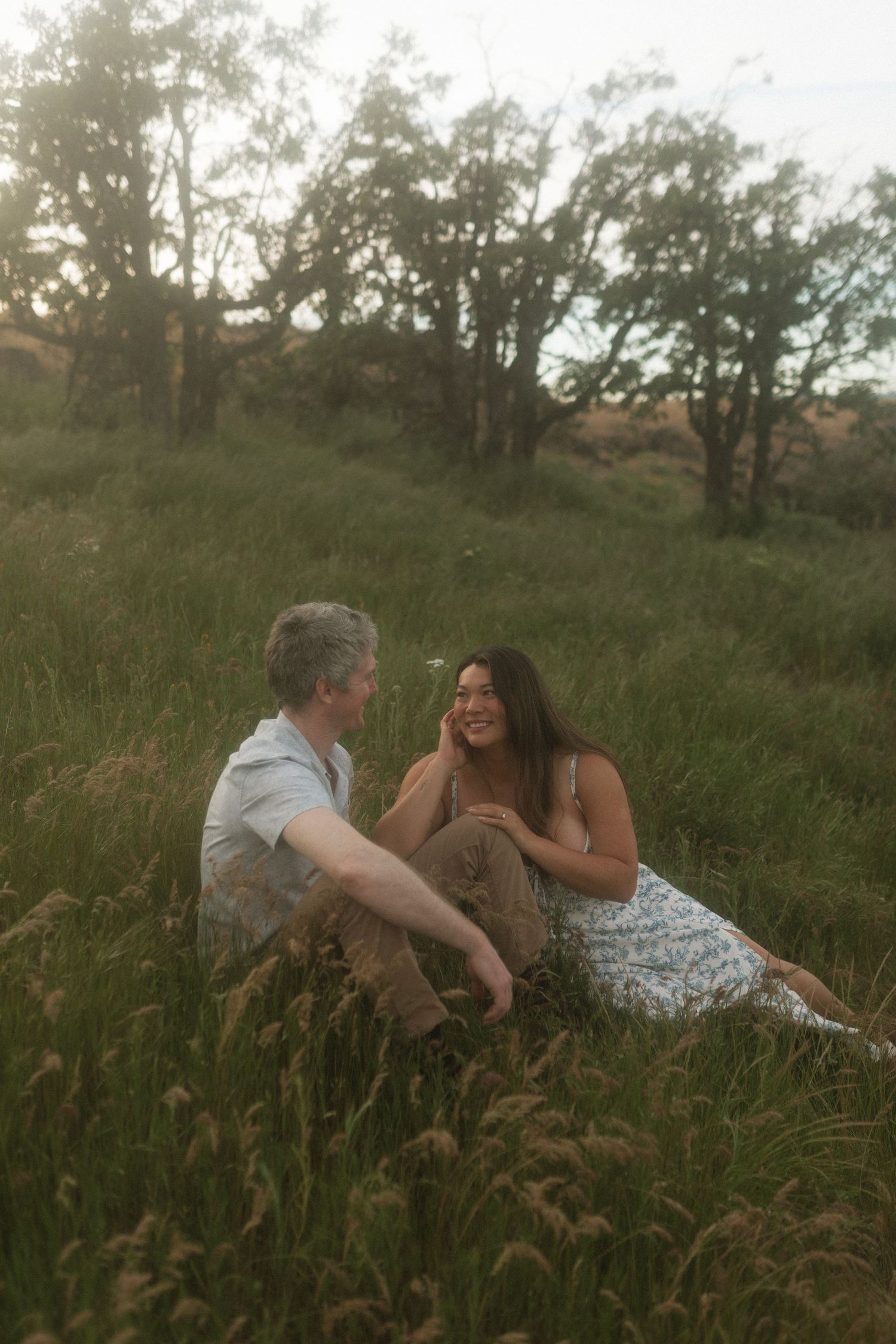 Couple sitting together in tall grass at sunset, smiling and sharing a tender romantic moment.