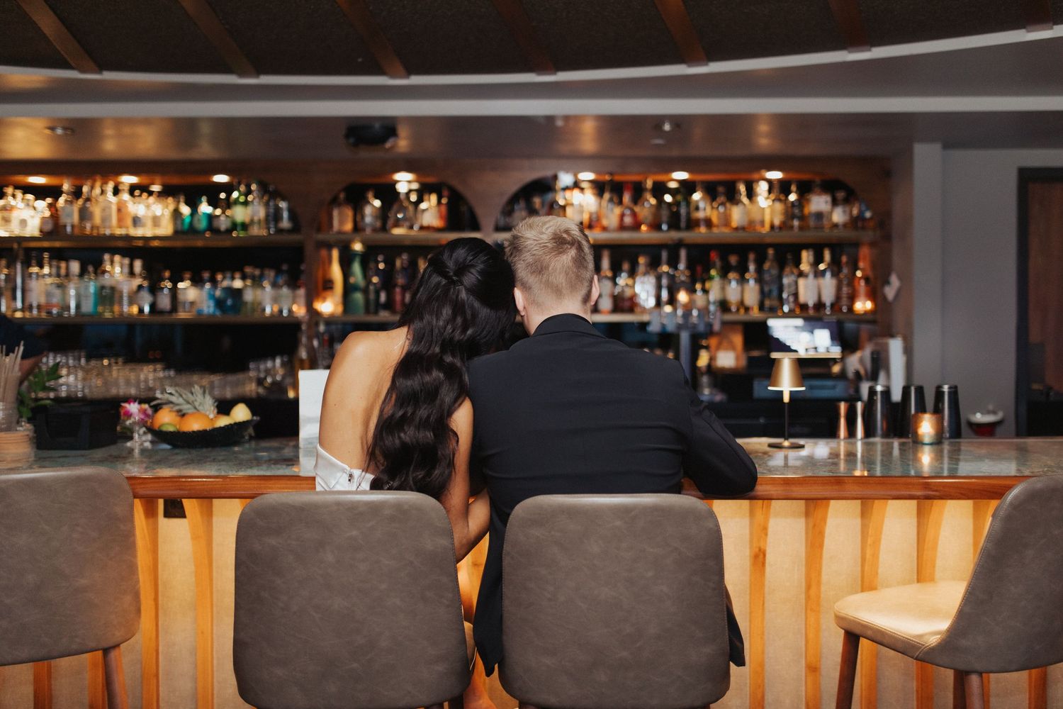 Couple sitting together at a luxurious hotel bar viewed from behind, surrounded by bottles and warm lighting.