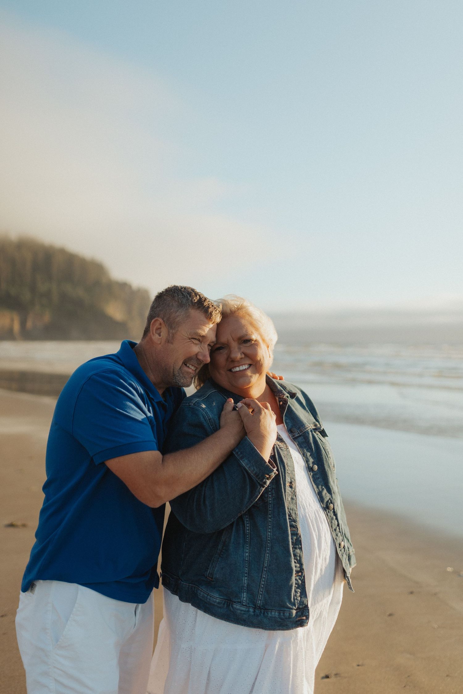 Affectionate mature couple embracing on a beach at golden hour with ocean and cliffs behind them.