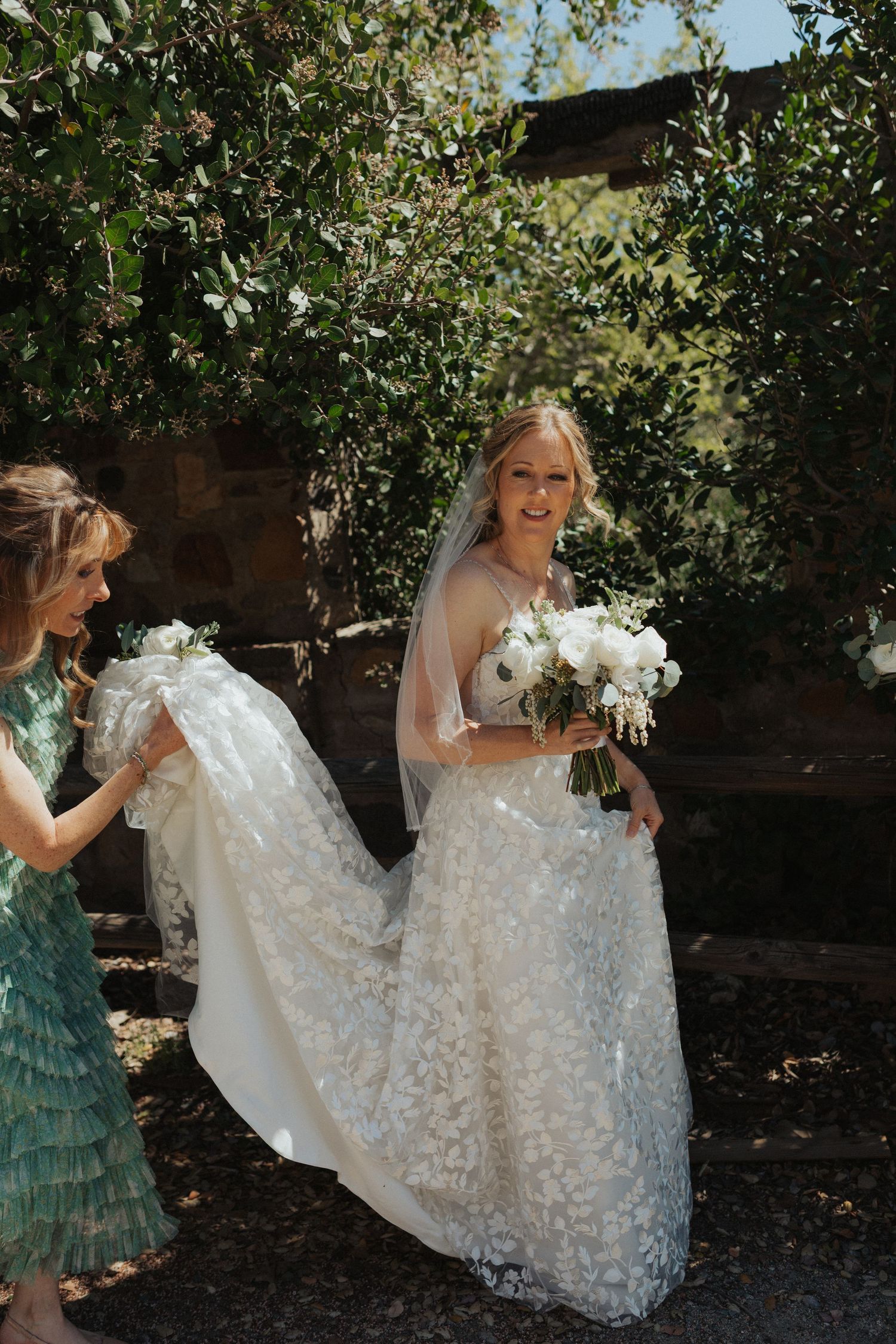 A smiling bride in a floral lace gown holds a white bouquet as a bridesmaid helps carry her train outdoors.