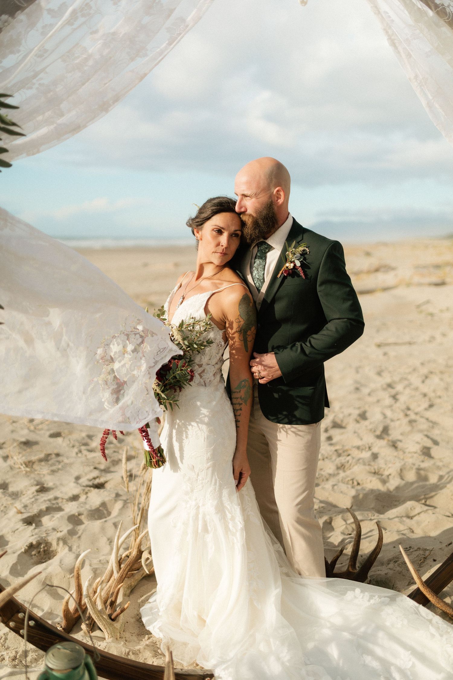 Bride and groom embrace under a draped arch on a sandy beach with a flowing veil.
