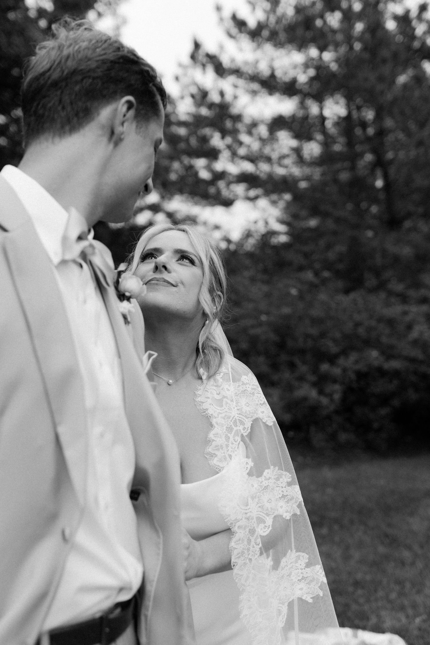 Black and white portrait of bride gazing up at groom surrounded by lush green trees outdoors.