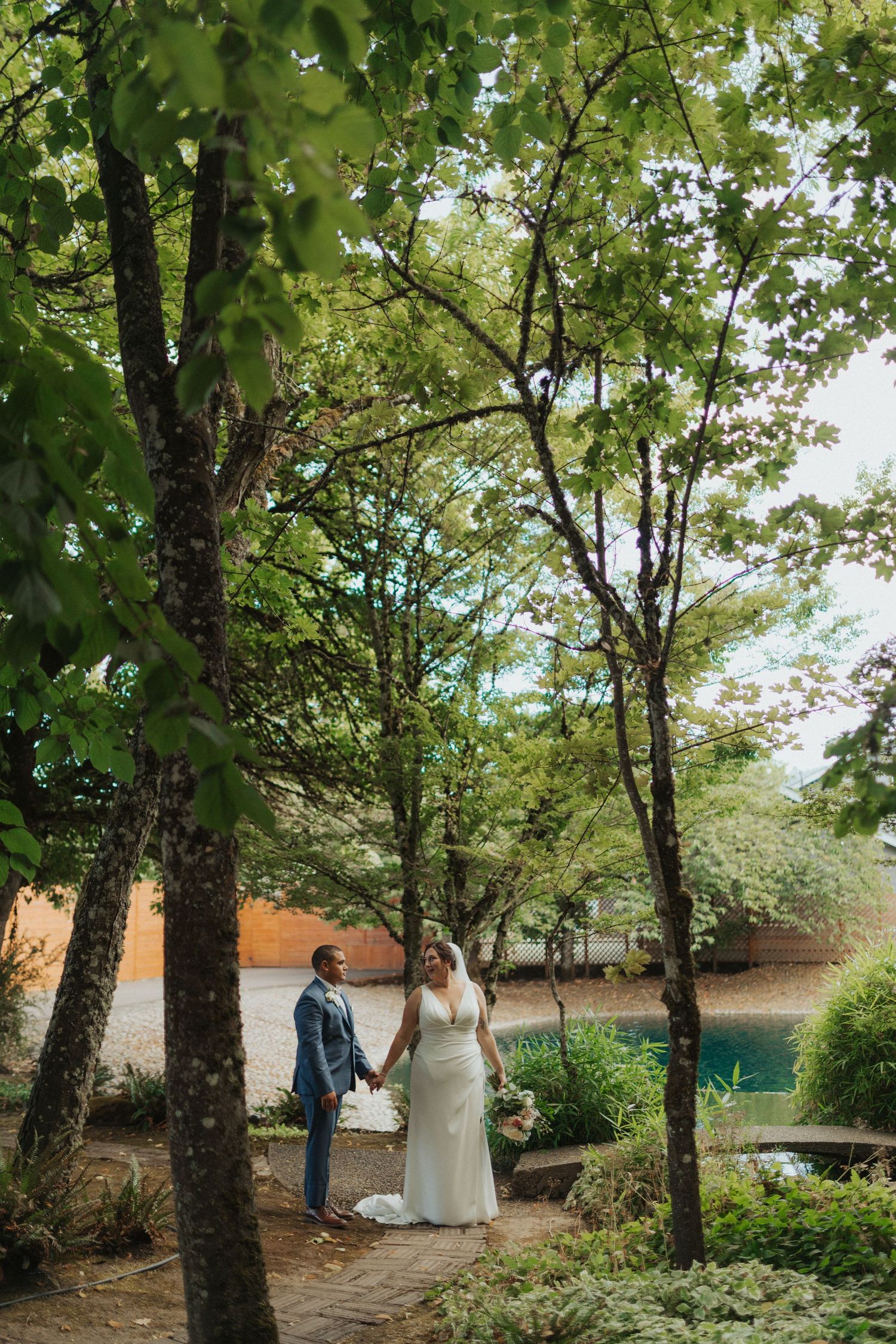 Wedding couple holds hands among tall green trees with a tranquil pond visible in the background.