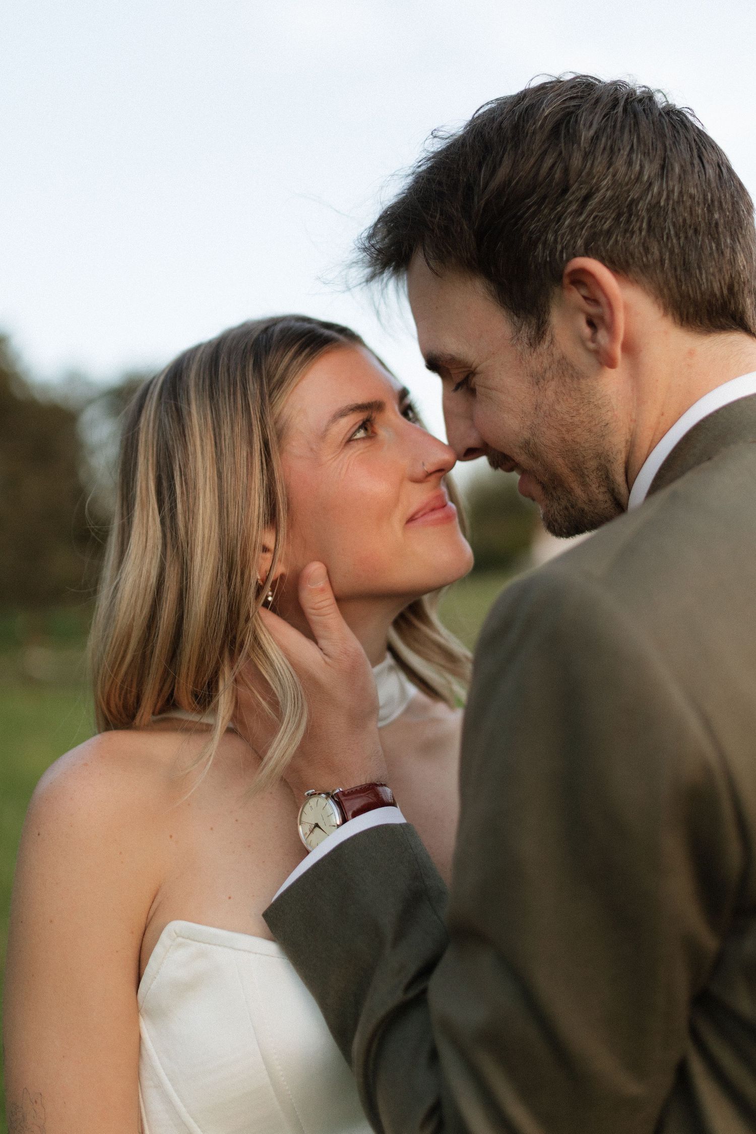 A bride and groom share an intimate moment, almost kissing, as the groom tenderly cups her face outdoors.