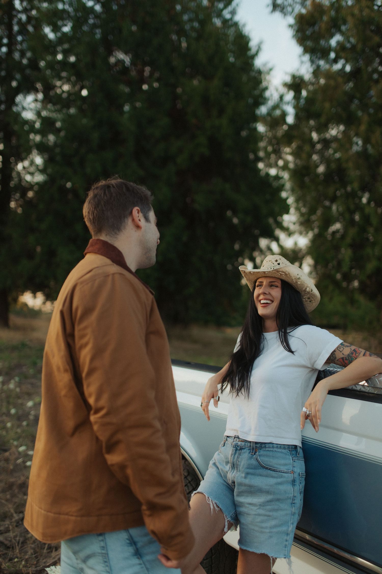 A laughing woman in a cowboy hat leans on a vintage car with her partner.