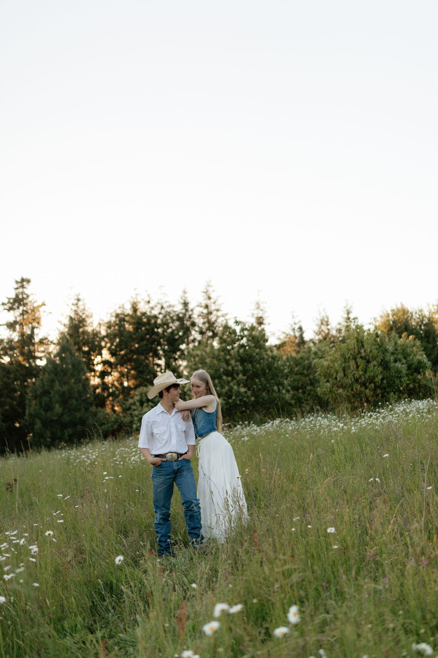 Couple embracing in a wildflower field at golden hour, man wearing cowboy hat and white shirt.