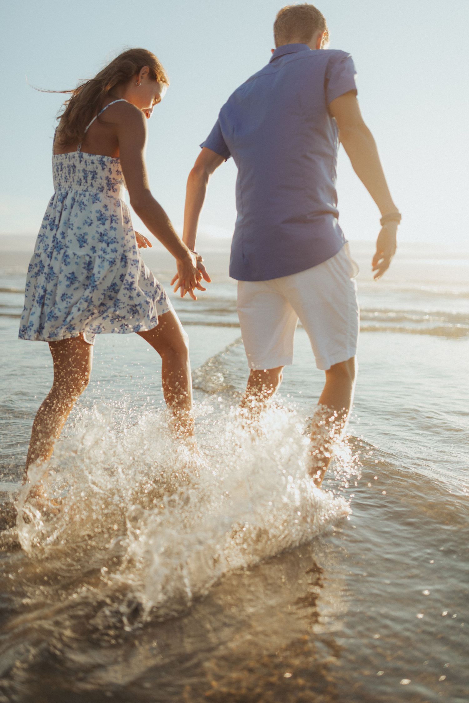 Couple splashing in ocean waves at sunset, woman in floral dress and man in blue shirt holding hands.