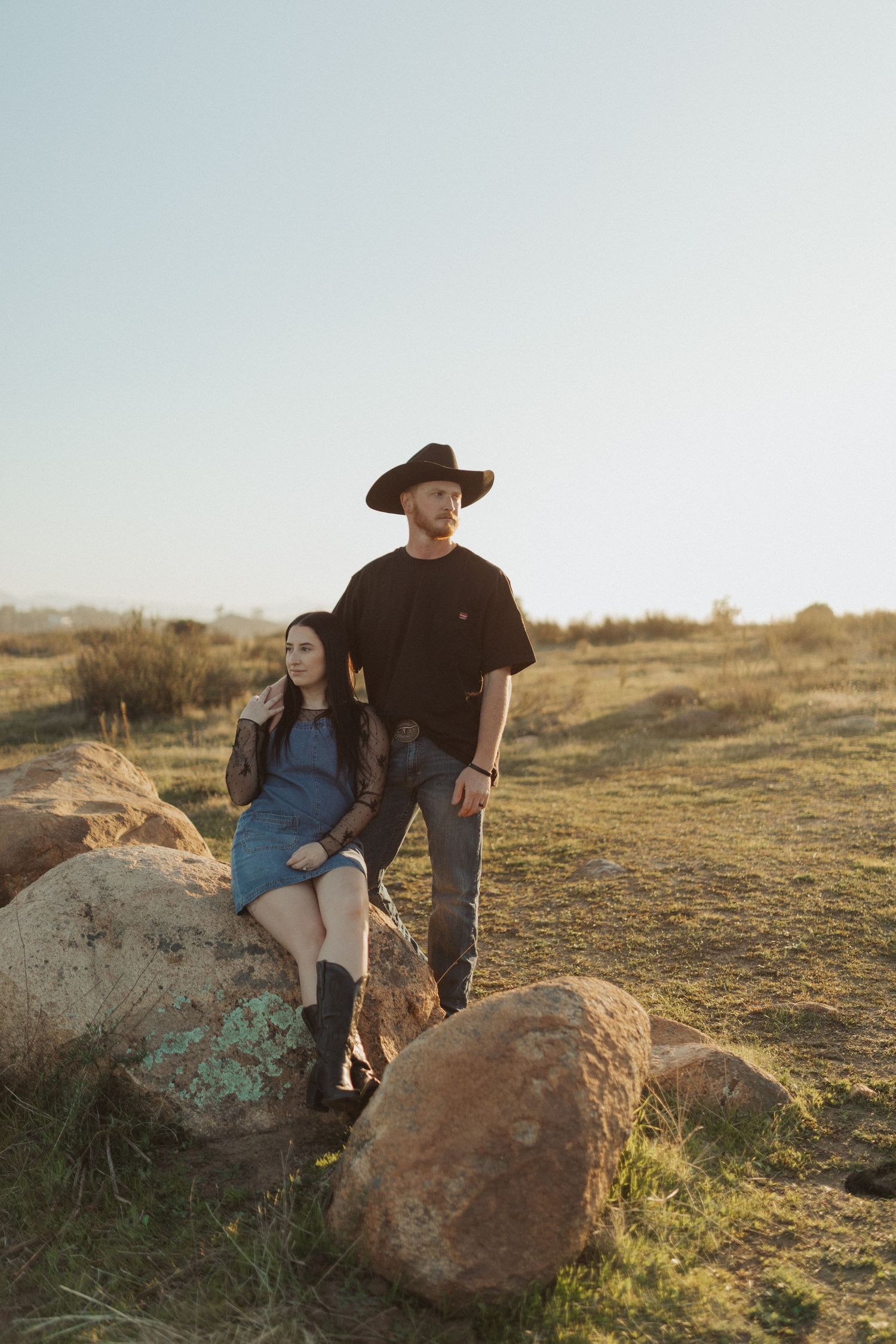 Couple posing on rocks in golden hour sunlight, man in black shirt and cowboy hat with woman in denim dress.