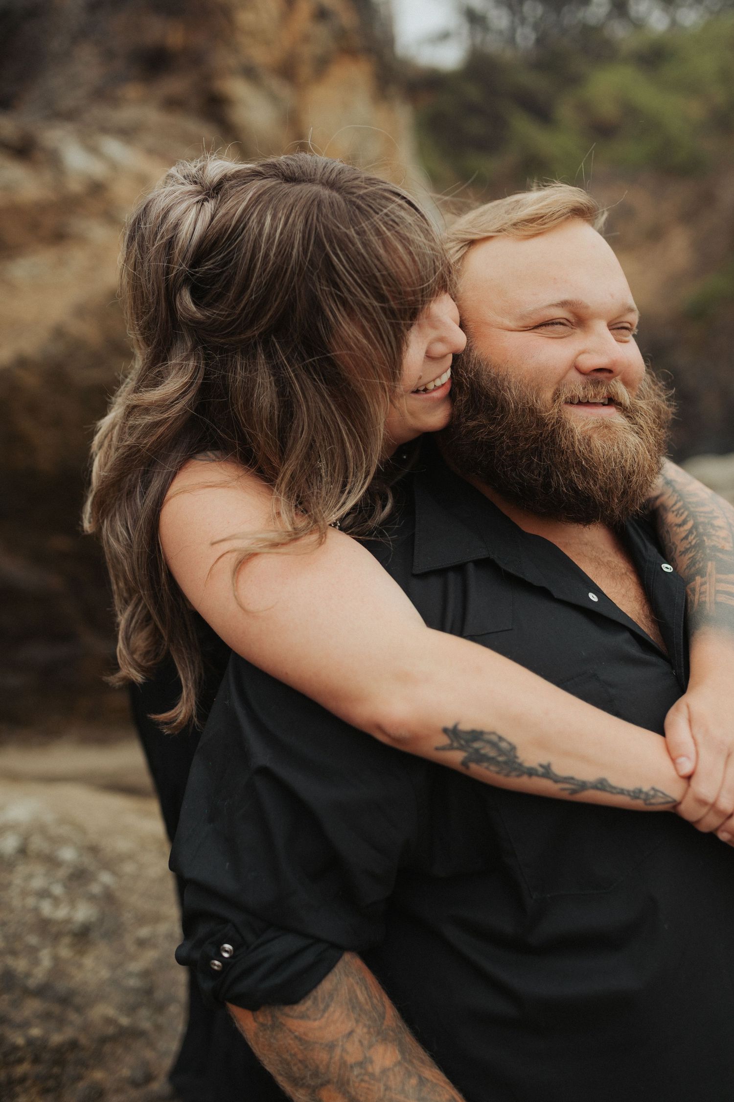 Bearded man smiling as tattooed woman hugs him from behind near rocky coastal cliffs.
