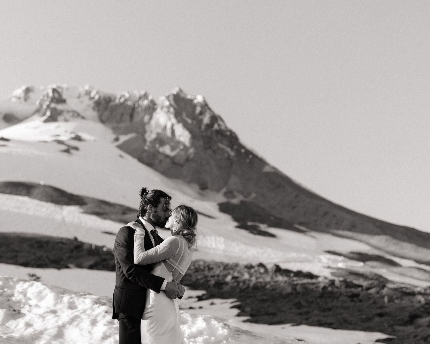 Black and white photo of bride and groom embracing with a snowy mountain peak behind them.