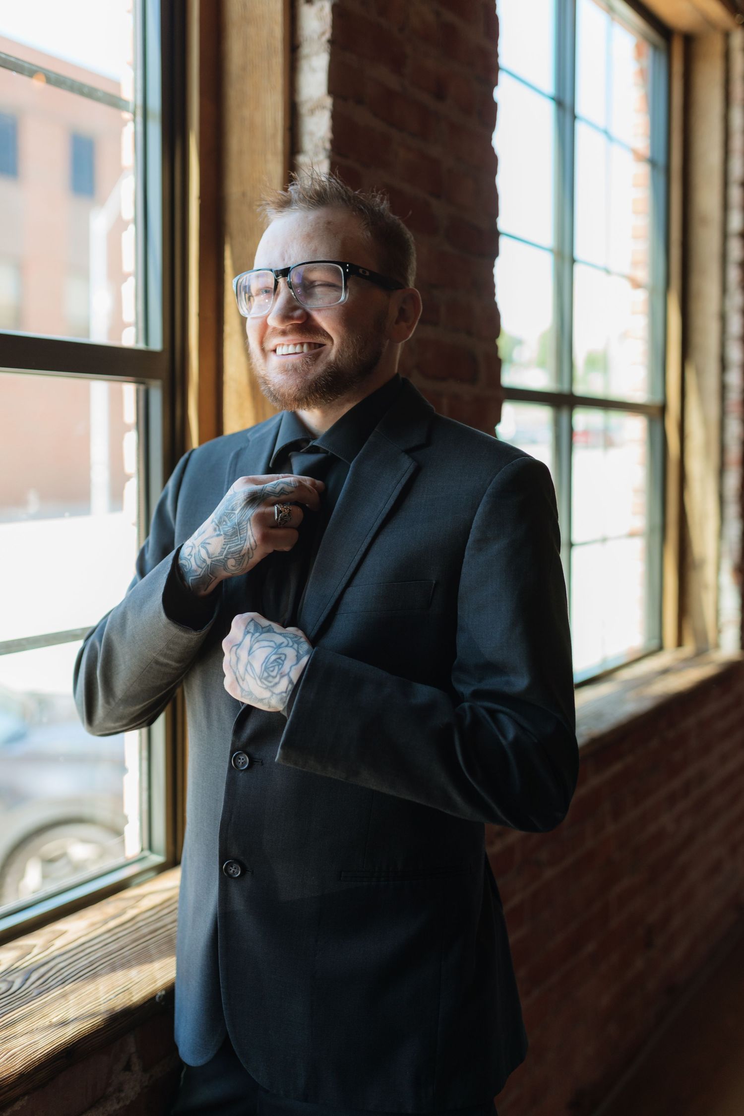 Smiling tattooed groom in black suit adjusting tie near large windows in a brick venue.