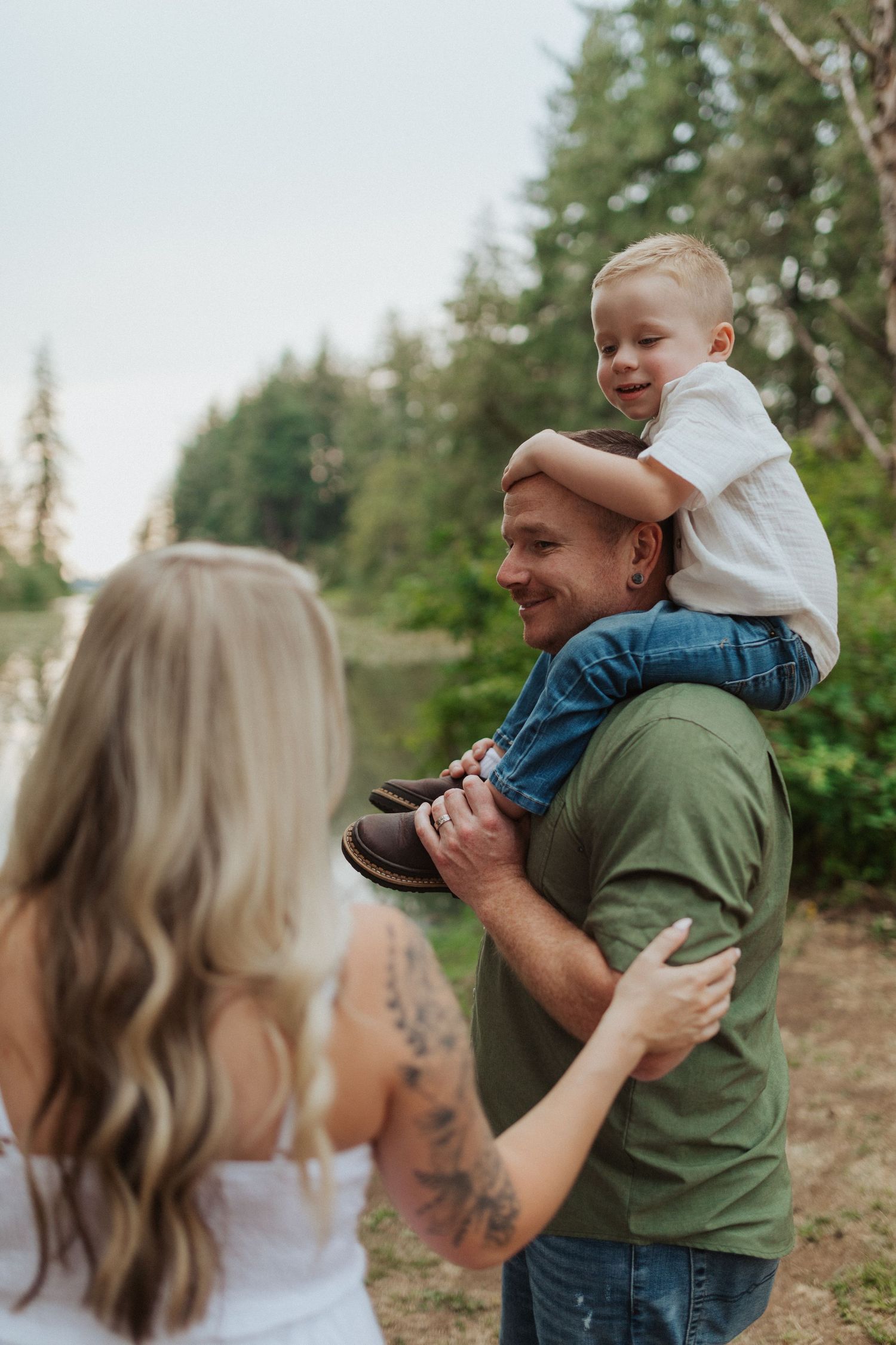 Laughing father carries young son on shoulders while mother watches in lush forest setting.