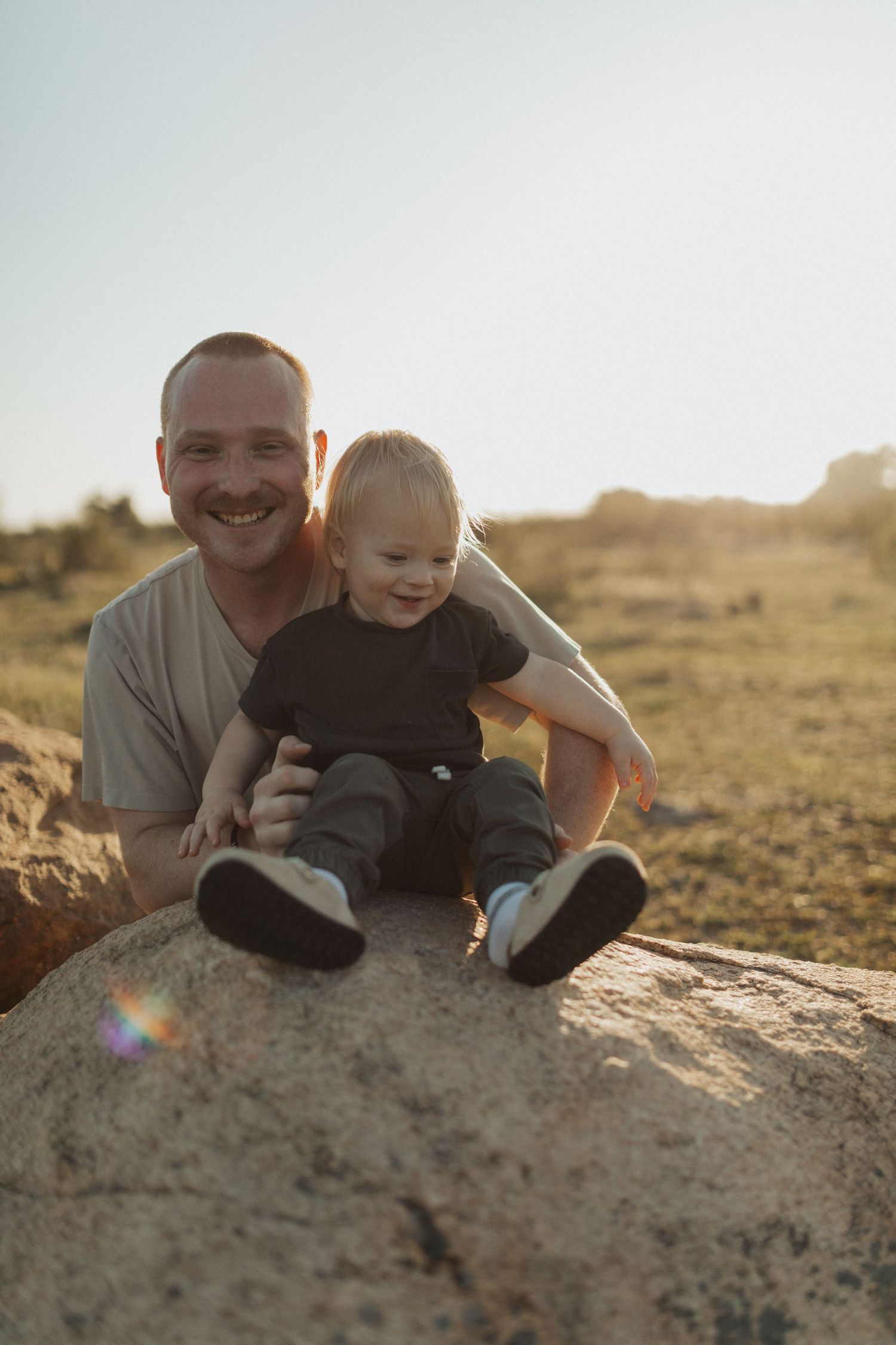 Smiling father sitting on boulder with happy toddler in black outfit during golden hour outdoors.